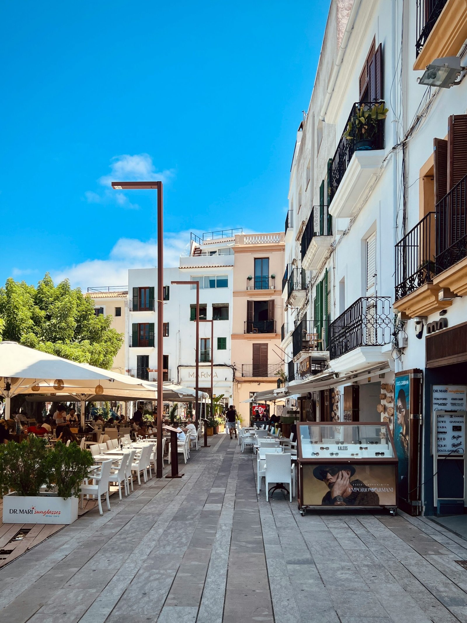 A sunlit pedestrian street in Ibiza Town with white-washed buildings, boutique shops, and outdoor cafes under a clear blue sky, capturing the high-end lifestyle of an Ibiza wellness retreat.