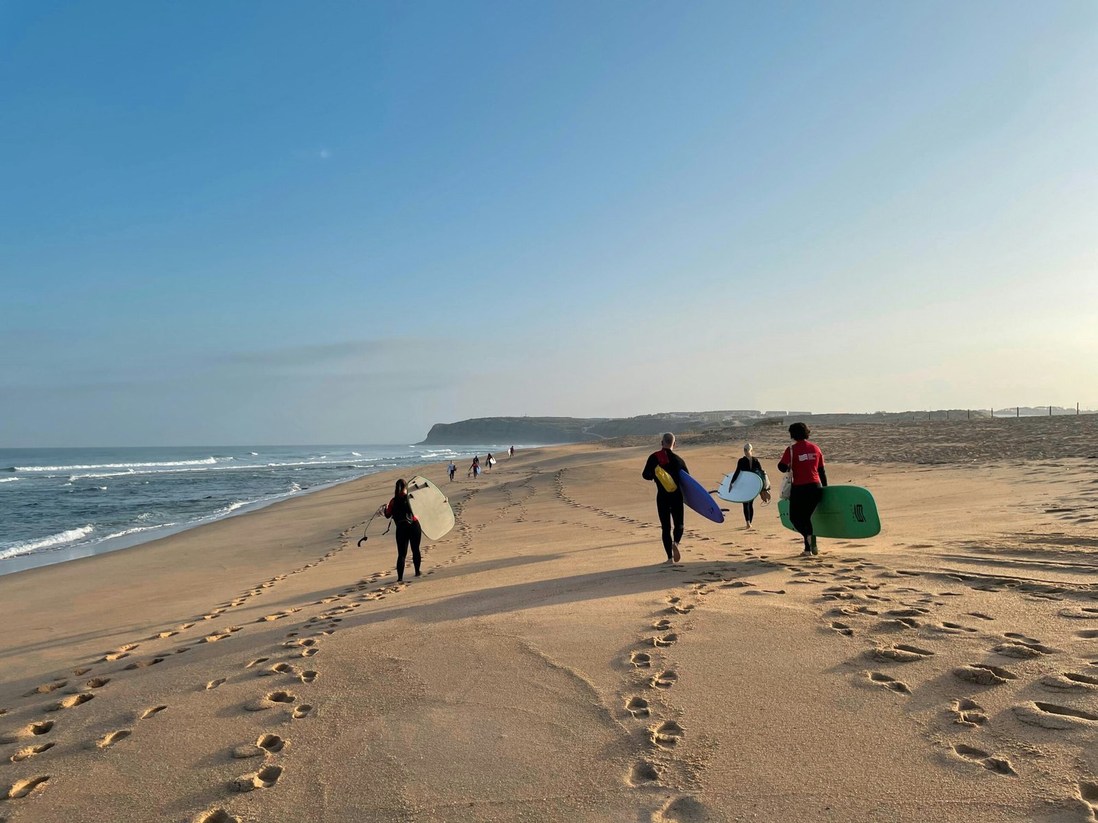 A group of surfers carrying their boards along a wide, sun-drenched beach in Portugal, capturing the active and communal spirit of a summer yoga and surf retreat.