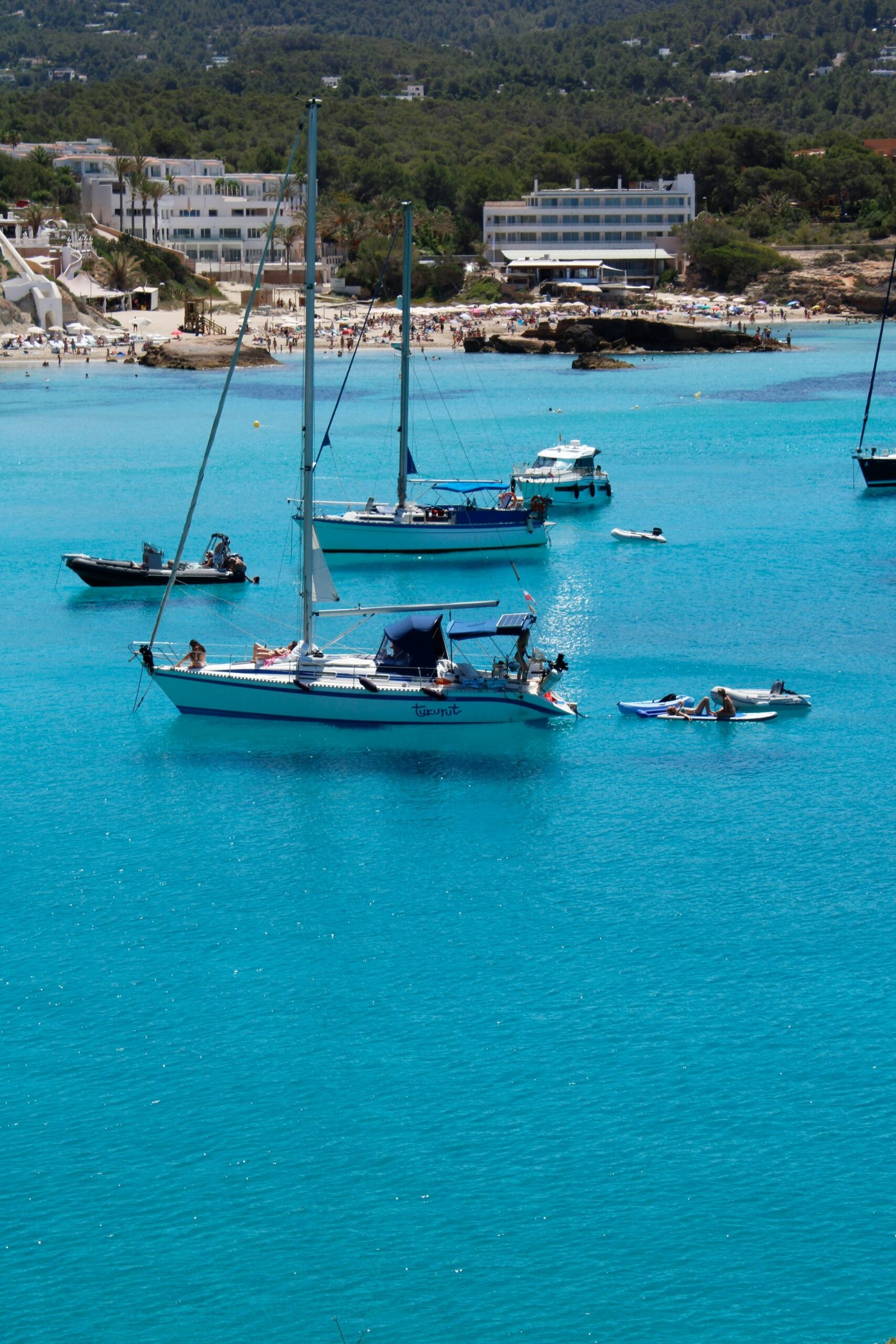 Several sailboats and small motorboats floating on the crystal-clear turquoise waters of an Ibiza bay, showing the peaceful Mediterranean lifestyle of a wellness retreat.