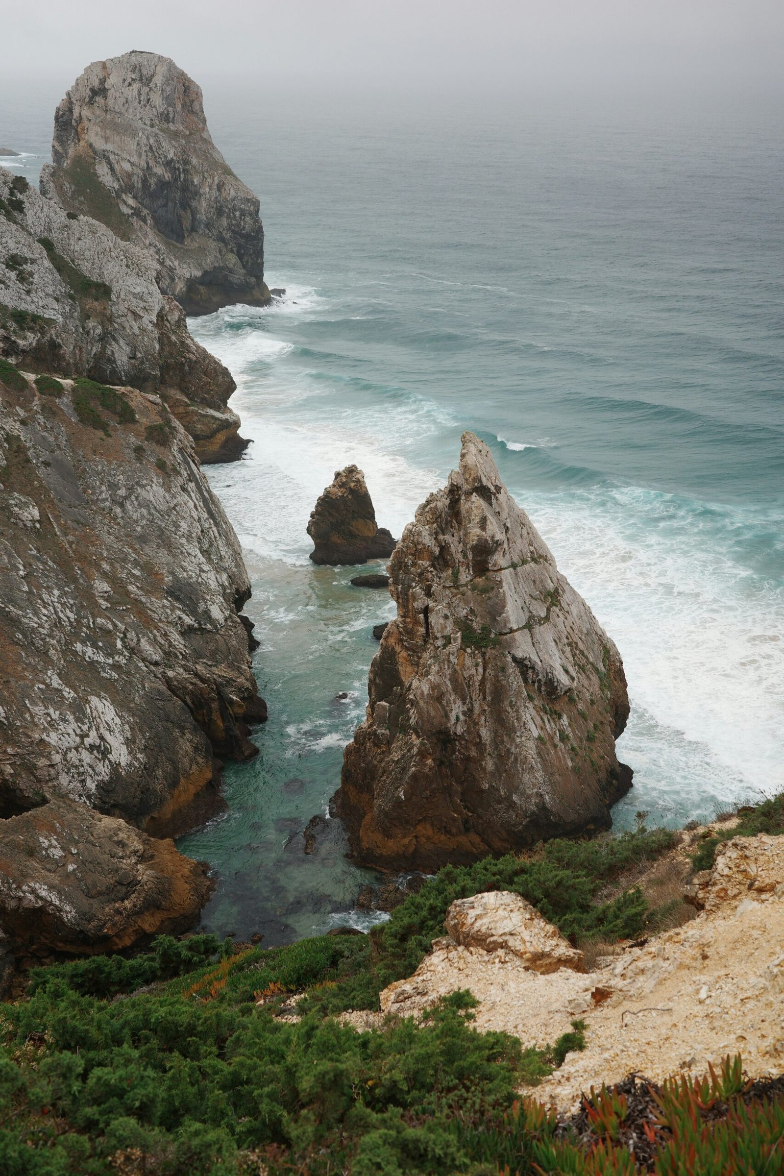 The dramatic, mist-covered sea stacks and limestone cliffs of the Sintra-Cascais coast, illustrating the wild and atmospheric 'Ocean Air' setting of yoga retreats near Lisbon.