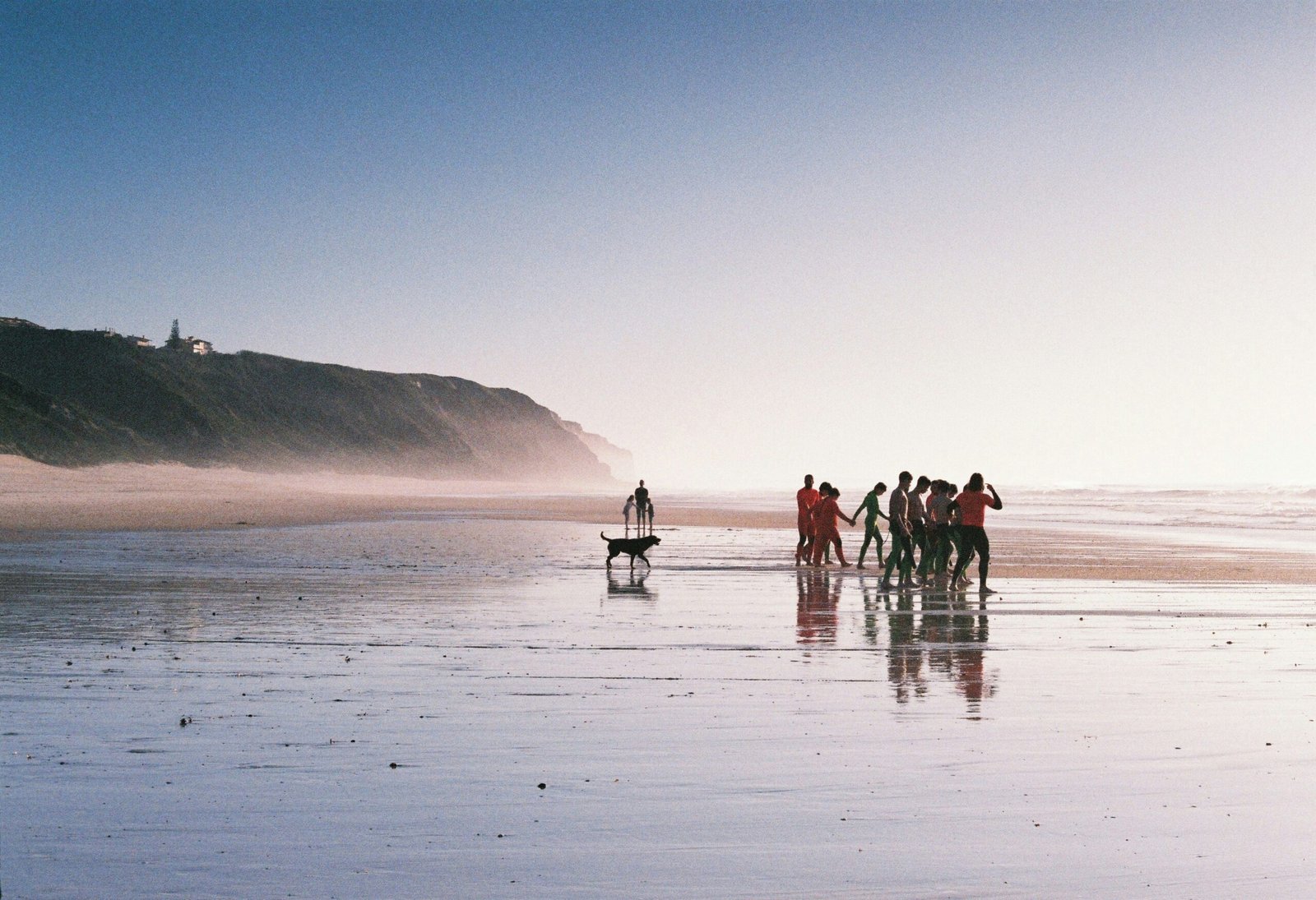 A group of yoga retreat participants in colorful wetsuits standing in a circle on a misty Ericeira beach at low tide, illustrating community and shared wellness experiences in Portugal.