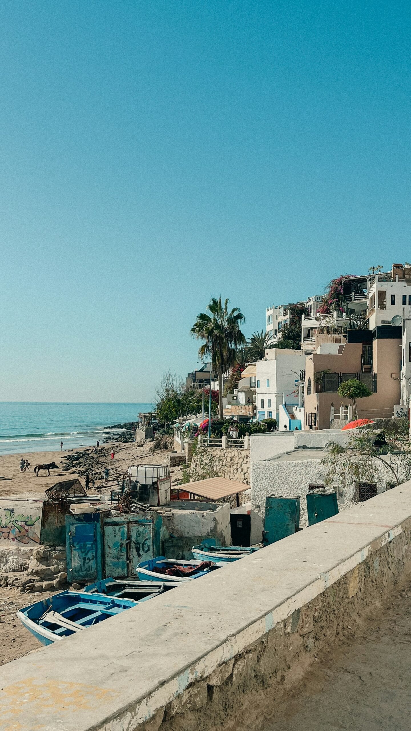 A bright coastal view of Taghazout, Morocco, featuring white-washed buildings on a cliff overlooking a sandy beach with blue fishing boats, ideal for a September surf and yoga retreat.
