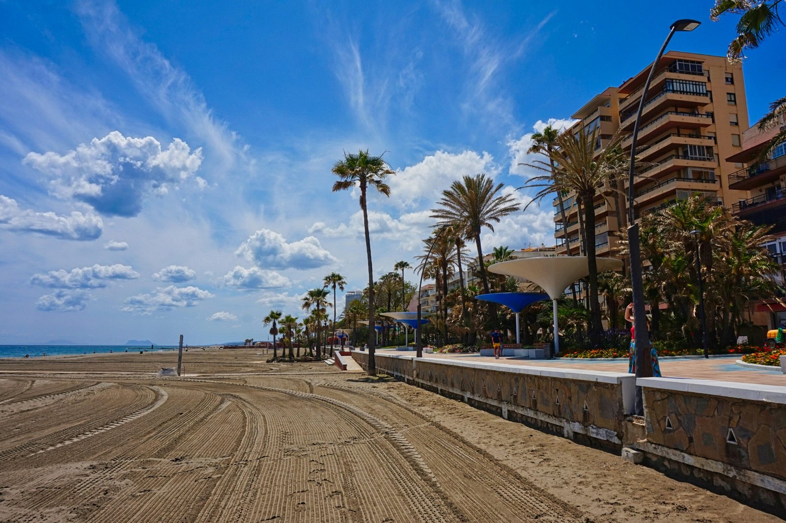 A wide, raked sandy beach in Spain under a bright blue sky with wispy clouds, featuring a palm-lined promenade and coastal architecture perfect for a restorative yoga retreat."