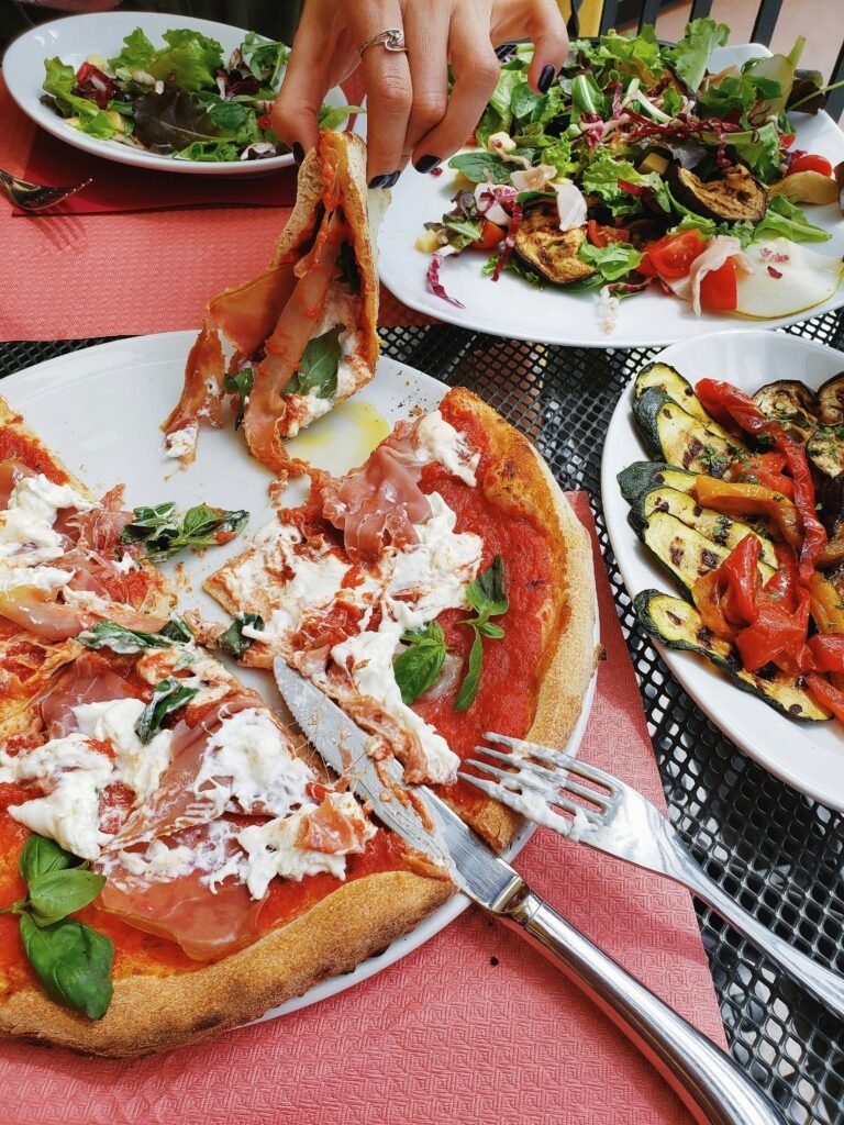 A vibrant table spread of Mediterranean food including a fresh burrata and prosciutto pizza, grilled vegetables, and seasonal salads, illustrating the art of slow nourishment.