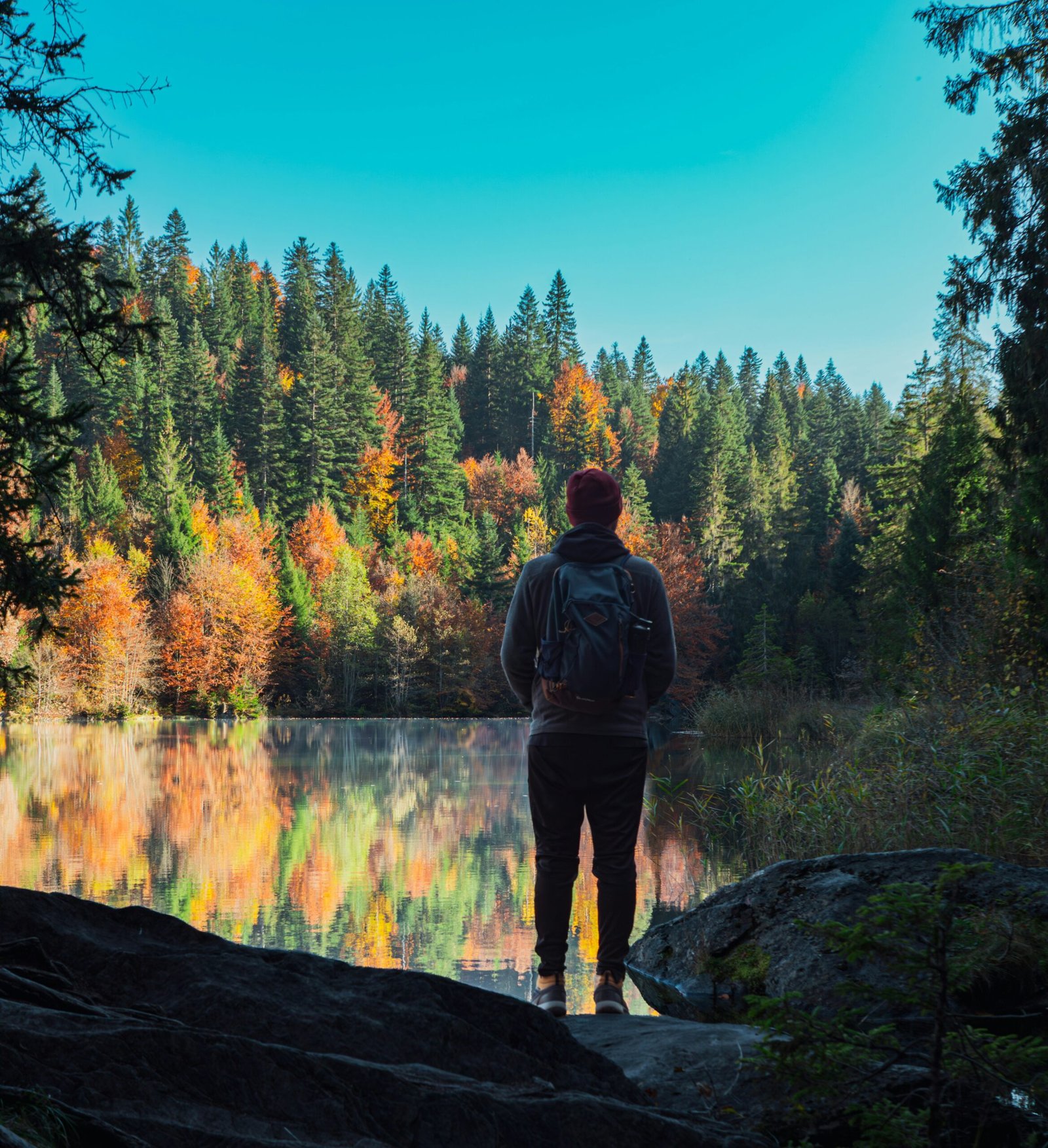 A hiker with a backpack standing before a still lake reflecting colorful autumn trees, illustrating the quiet introspection and deep nature immersion of a boutique wellness retreat.