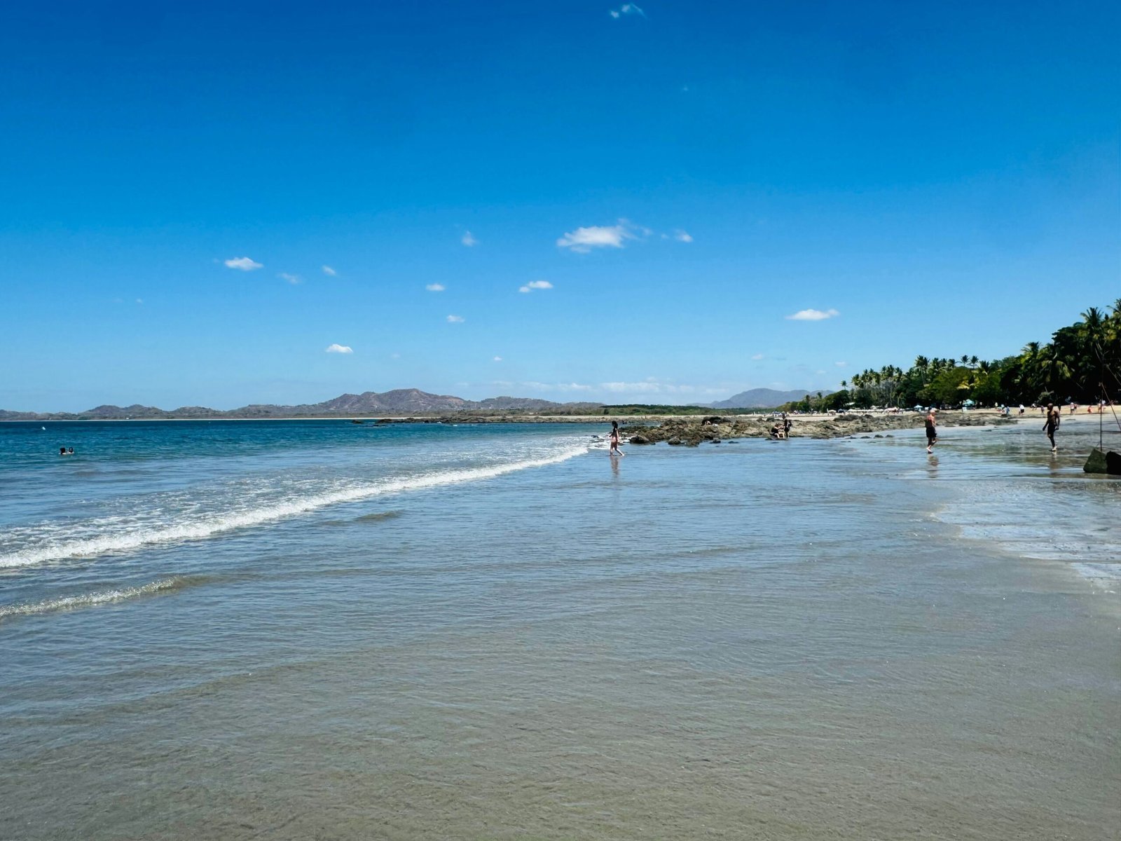 Sunny beach in Tamarindo, Costa Rica, with calm waves, golden sand, and hazy hills along the shoreline.