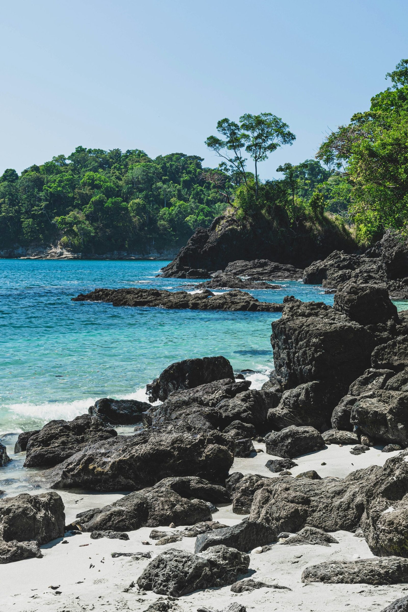 Rocky tropical coastline at Manuel Antonio Beach, Costa Rica, with green trees above dark volcanic cliffs and bright blue ocean water.