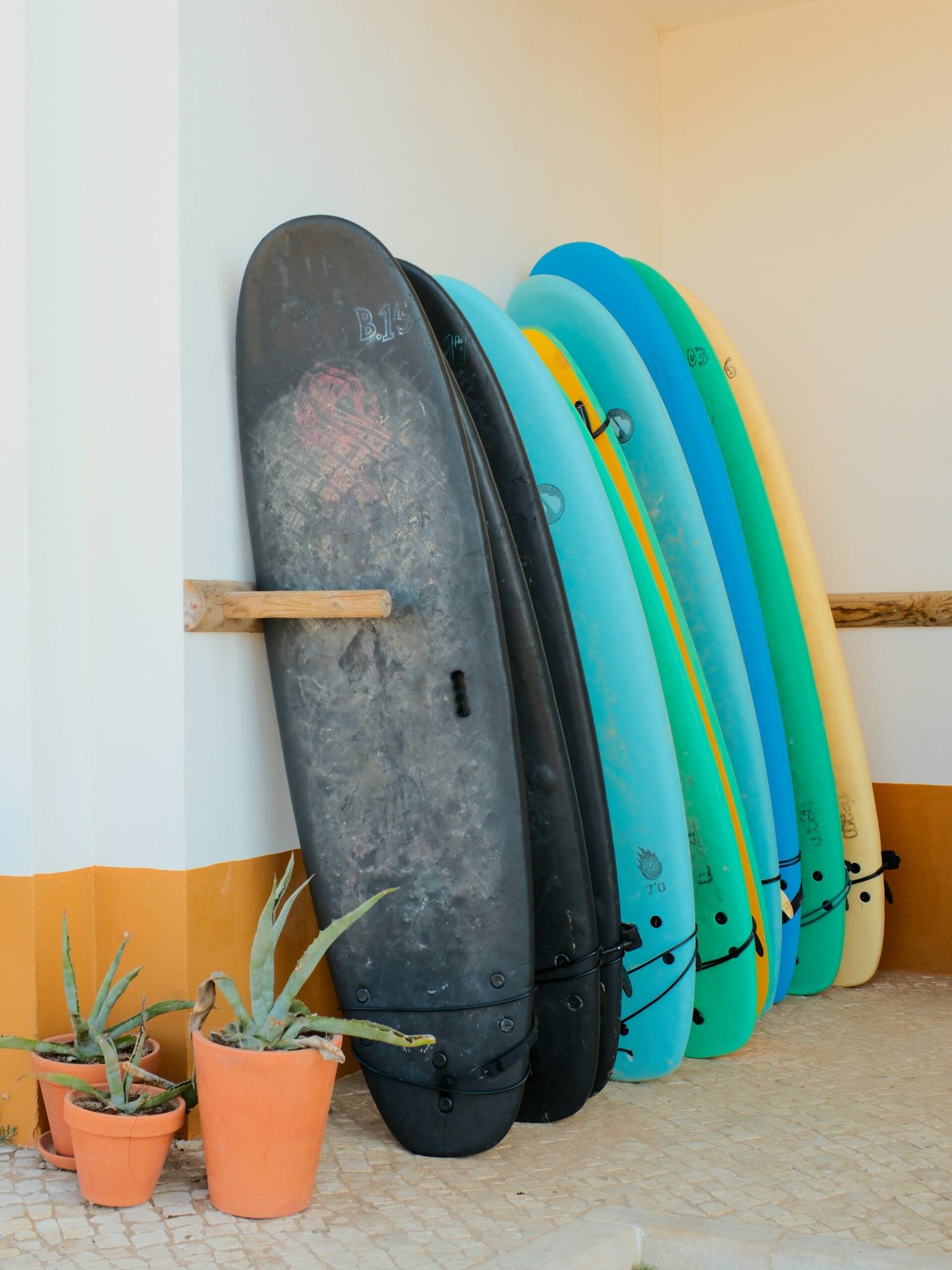 A row of colorful surfboards leaning against a white and yellow wall next to potted aloe vera plants at a Portuguese surf and yoga retreat.