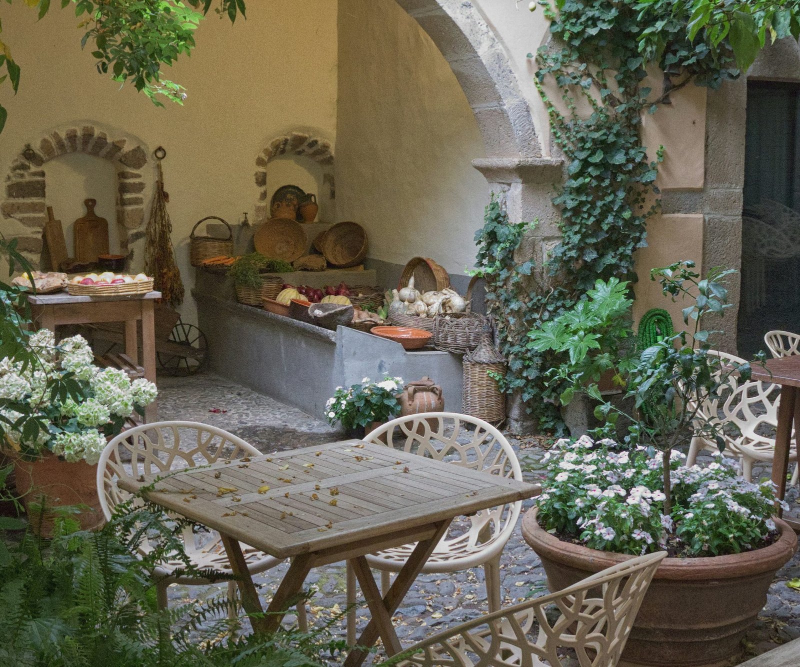 A rustic stone courtyard with an outdoor wooden dining table, fresh garden flowers, and baskets of produce, reflecting the peaceful atmosphere of slow nourishment in the Mediterranean.
