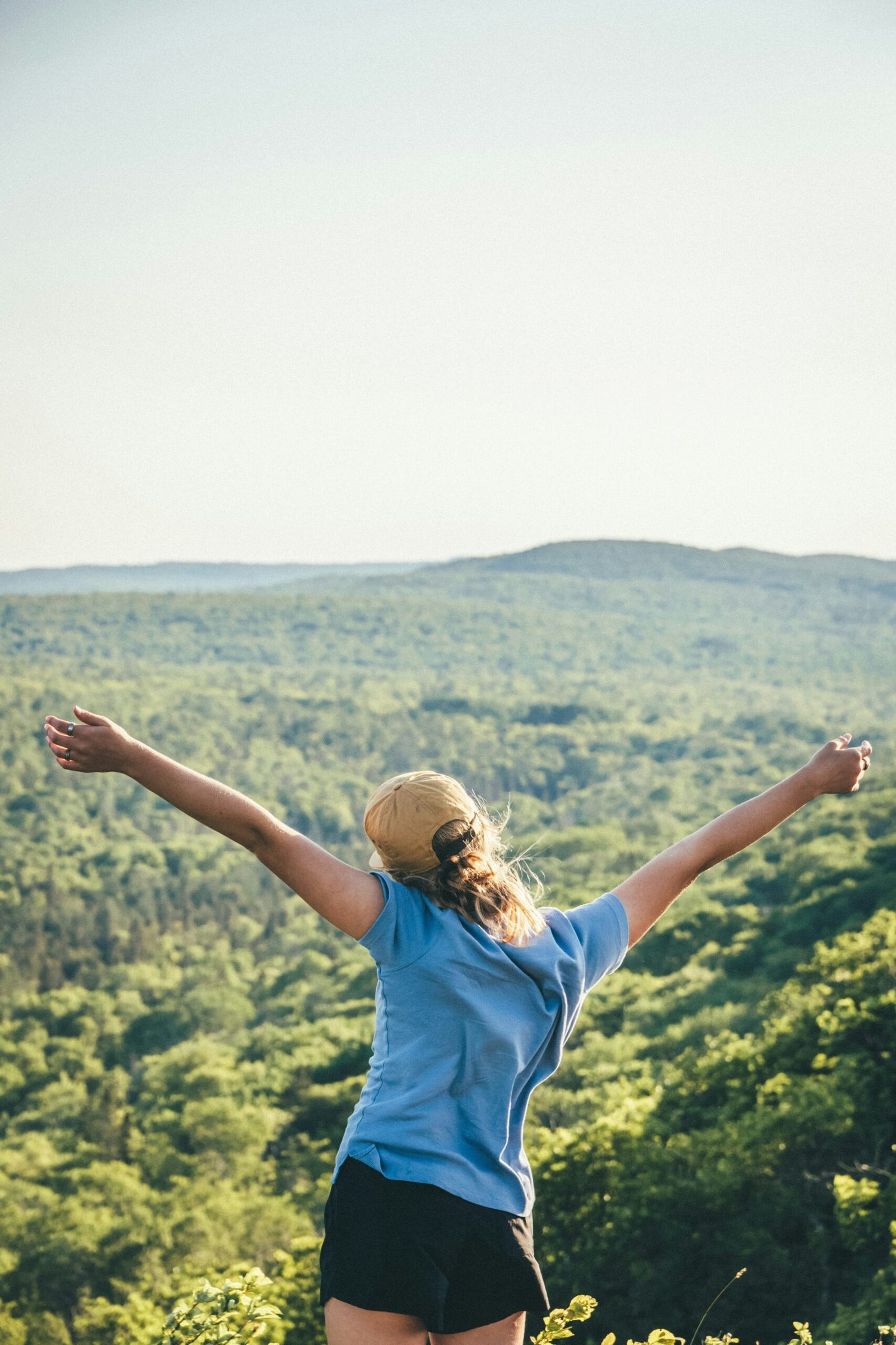 A young woman with arms open wide overlooking a vast green valley, symbolizing the sense of freedom and accomplishment felt after completing a multi-day nature retreat.