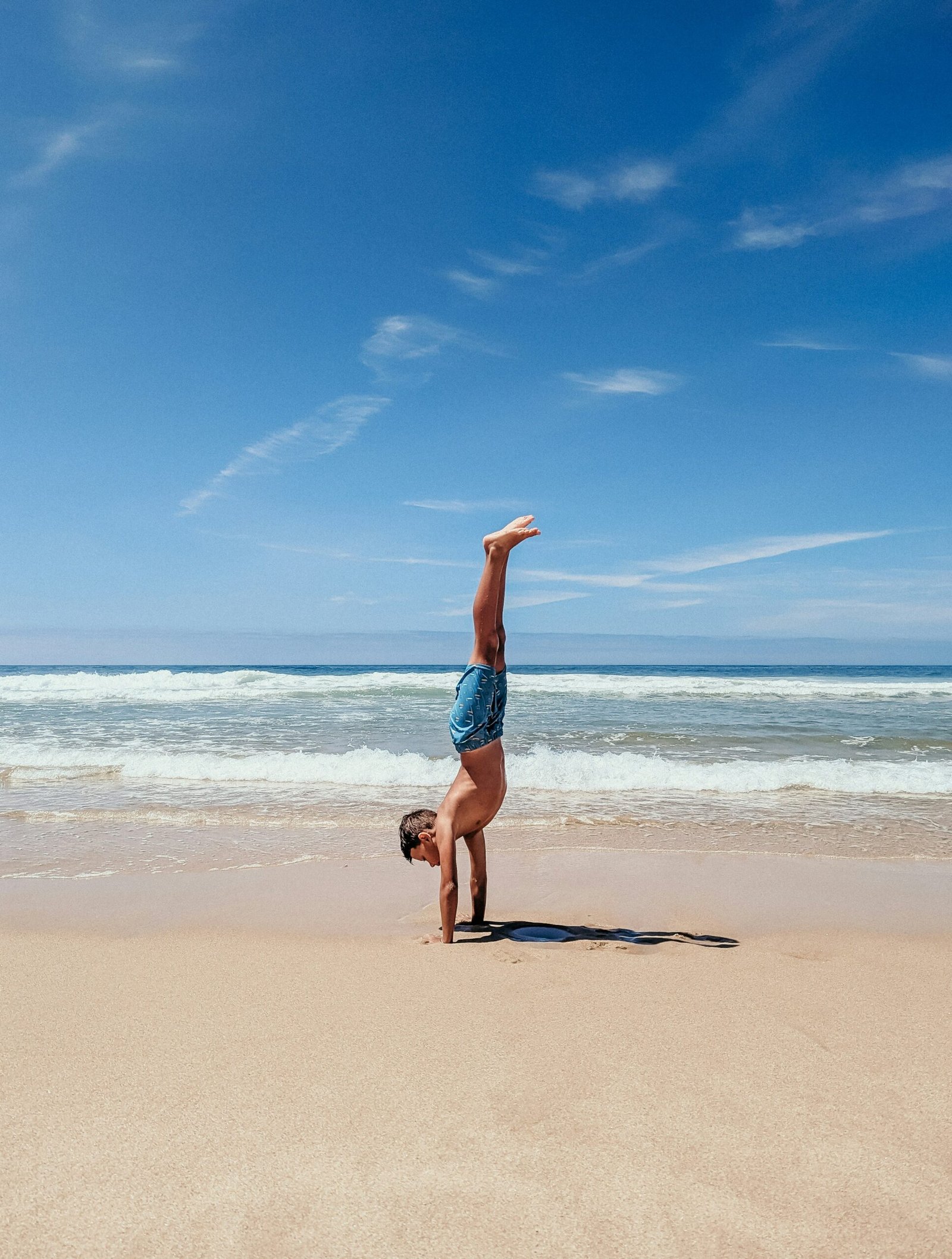 A young man performing a handstand on a wide, sandy beach under a clear blue sky, illustrating the energetic and youthful appeal of active yoga retreats in coastal Portugal.