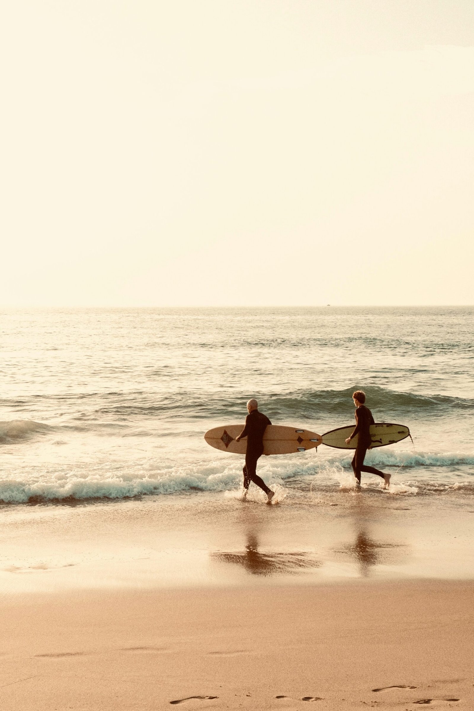 Two surfers carrying boards into the ocean at sunset near Lisbon, Portugal, highlighting the popular combination of surfing and yoga at wellness retreats in the Cascais and Ericeira regions.