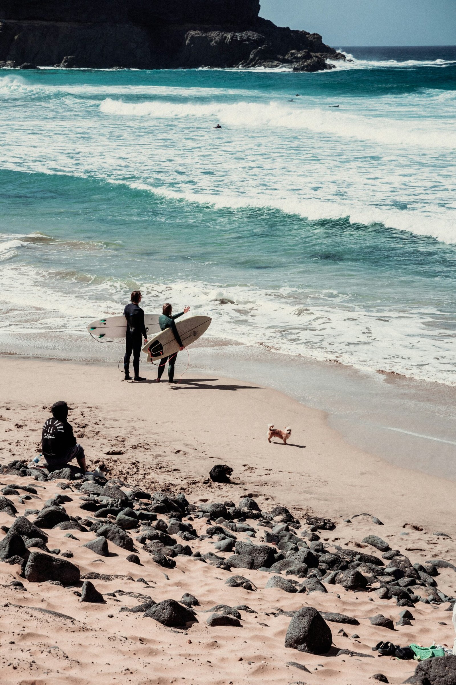 Two surfers in wetsuits with their boards watching the waves at a wide Atlantic beach in Spain, capturing the energy of a combined surf and yoga retreat.