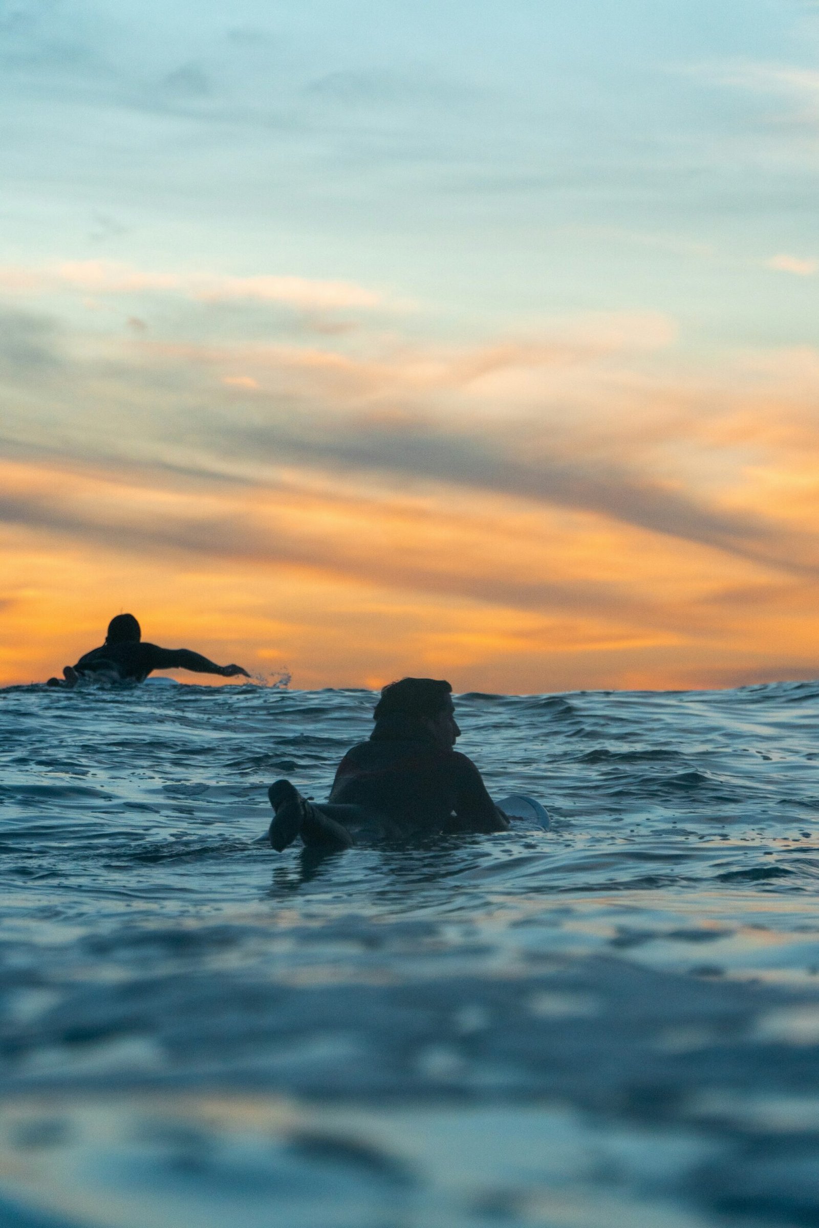 Two surfers paddling through the ocean at sunset in Ericeira, Portugal, capturing the meditative 'stillness' and connection to nature found during a surf and yoga retreat.