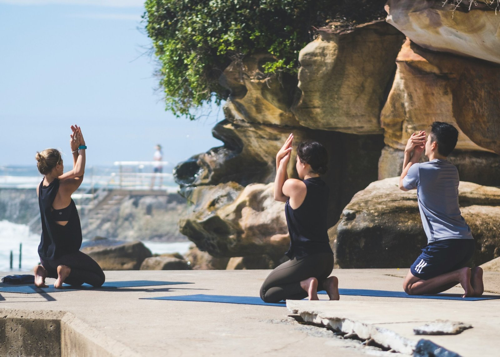 Three people practicing eagle pose during an outdoor yoga session on a stone platform by the Portuguese coast, highlighting the need for performance activewear and non-slip mats.