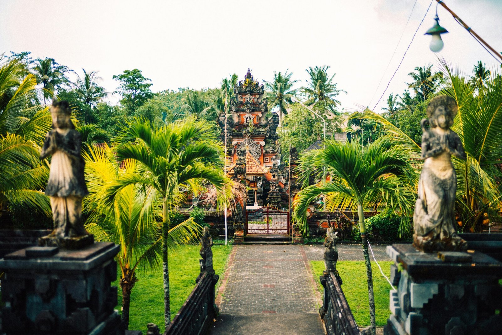 A traditional Balinese temple entrance with stone statues and palm trees under the bright Summer 2026 sun, representing the spiritual heart of a Bali yoga retreat.
