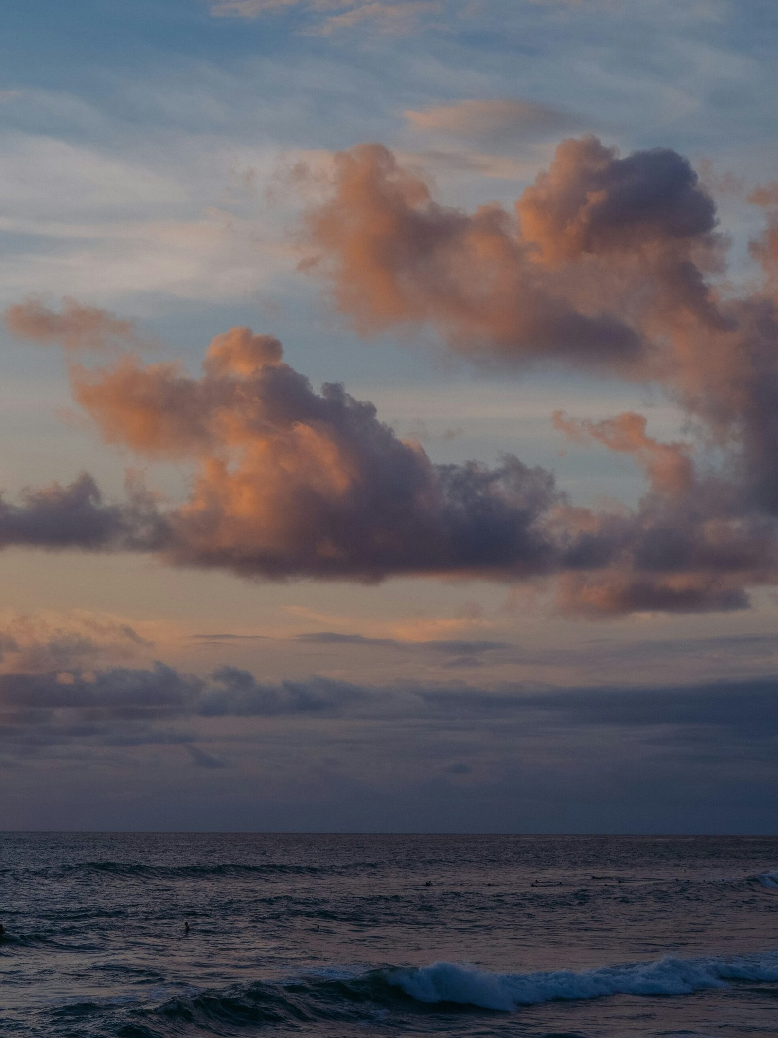 A serene twilight view of the Bali coastline with soft pink clouds and surfers in the water, capturing the tranquil atmosphere of a late-summer yoga retreat evening.