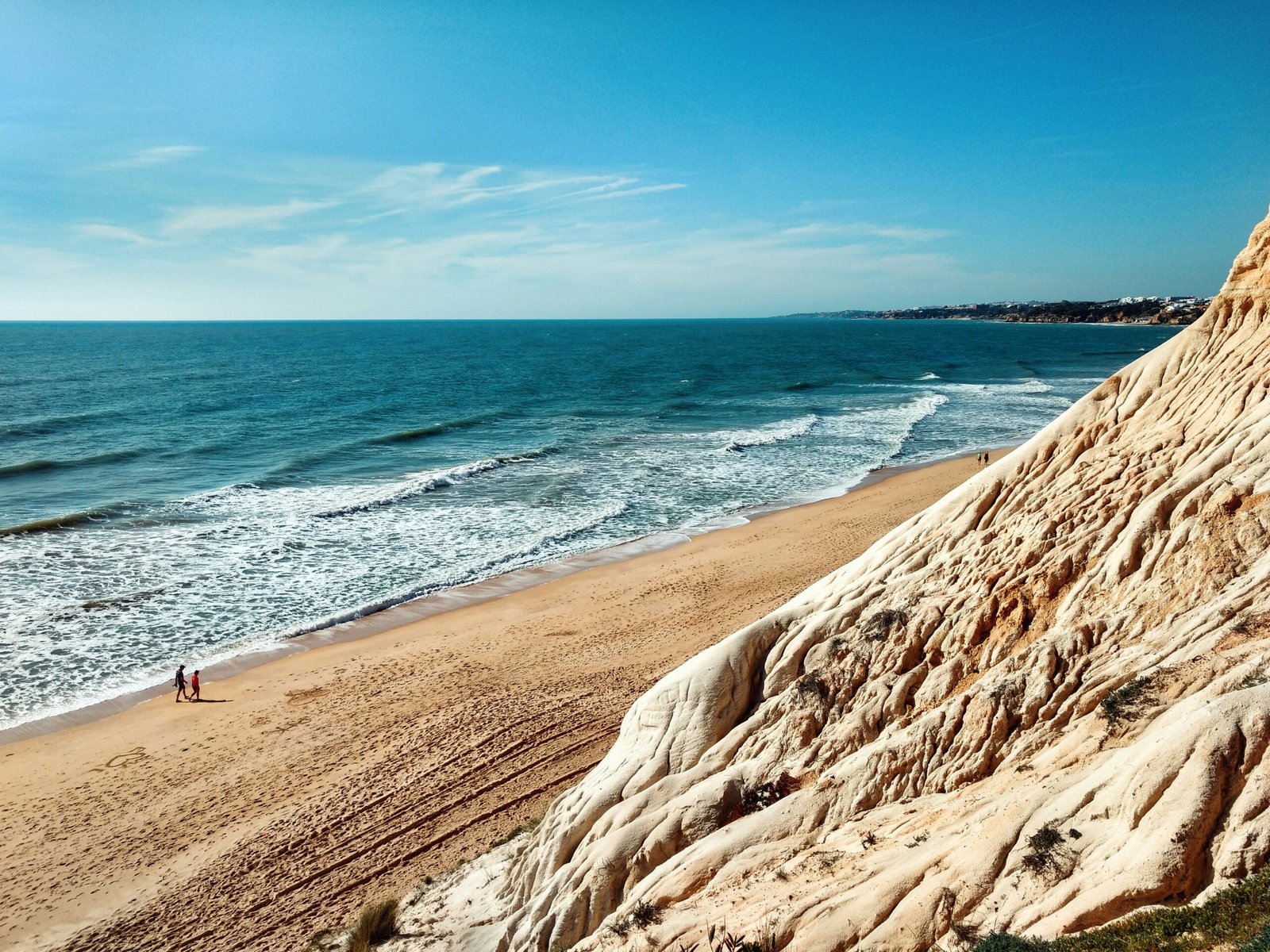 A vast, sun-drenched beach in the Algarve, Portugal, featuring towering orange-white sandstone cliffs and two people walking along the shoreline, ideal for an outdoor winter yoga and wellness retreat.