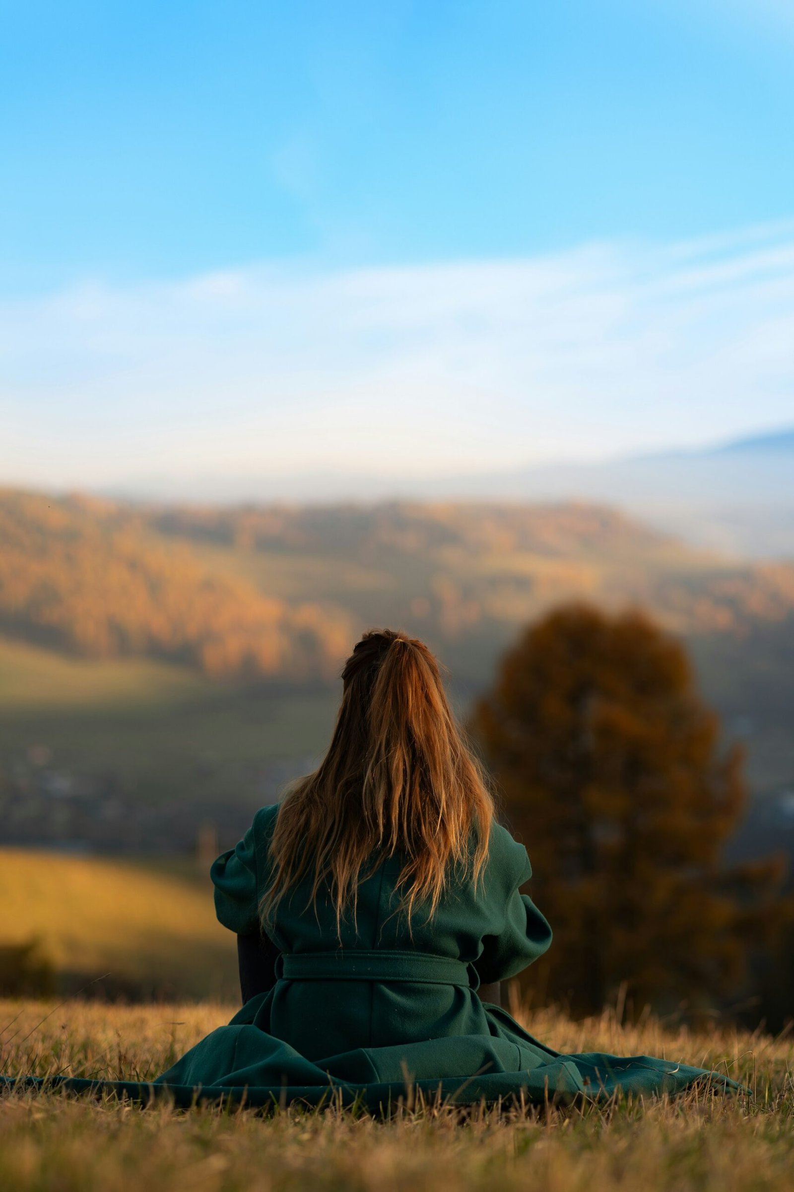A woman sitting on a grassy hilltop overlooking rolling autumn mountains, illustrating the long-term mental clarity and perspective gained from an extended wellness retreat.