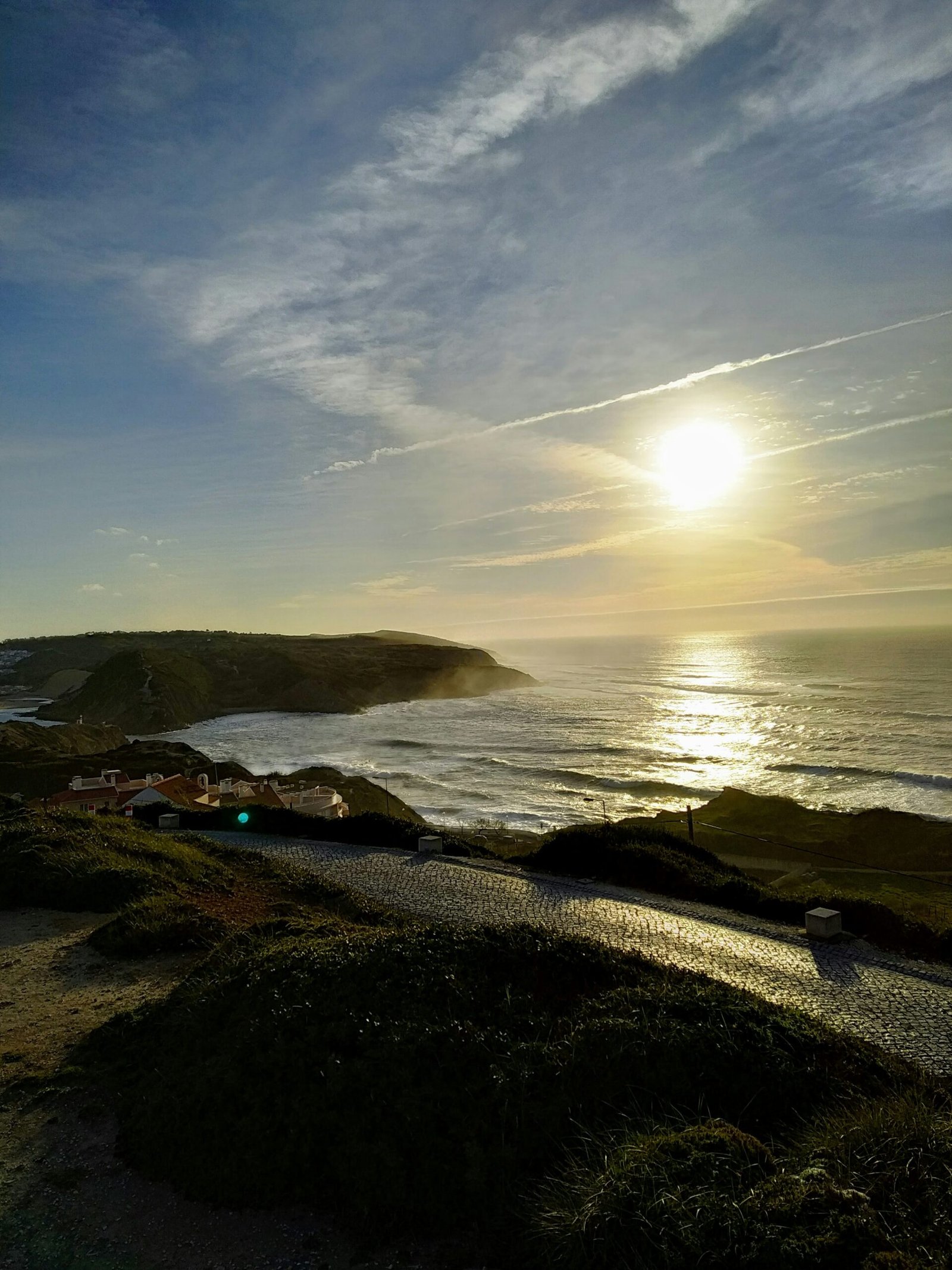 A wide coastal view of the Portuguese Atlantic at sunset during winter, featuring a cobblestone path winding through green dunes toward a misty bay and crashing waves.