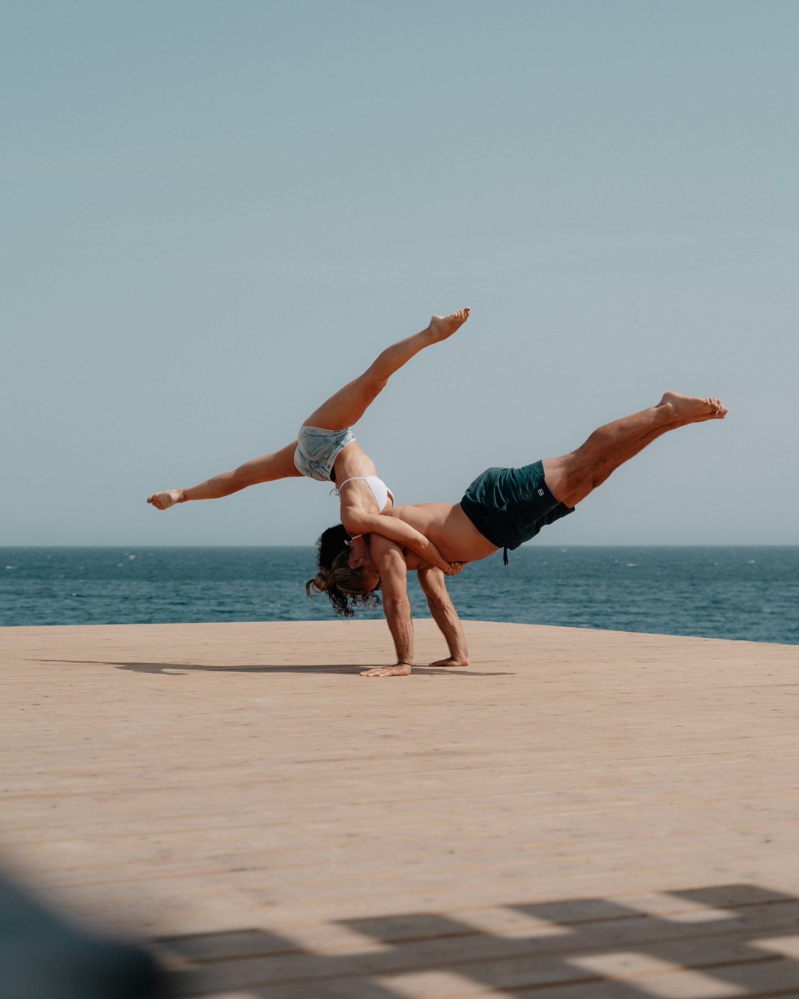 A male and female practitioner performing an advanced acro-yoga balance pose on a wooden deck overlooking the deep blue sea in Spain, capturing the peak experience of a surf and yoga retreat.
