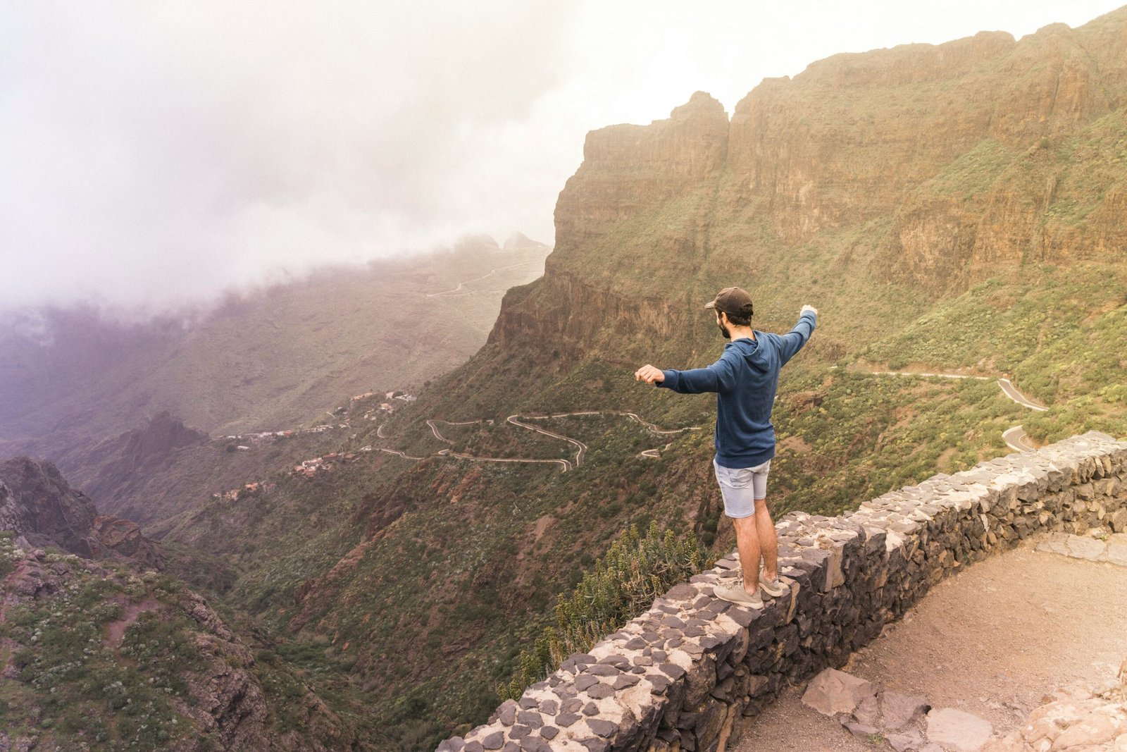 A man standing with open arms on a stone wall overlooking a dramatic mountain valley in the Canary Islands, representing the sense of freedom and expansion found at high-altitude yoga retreats in Spain.