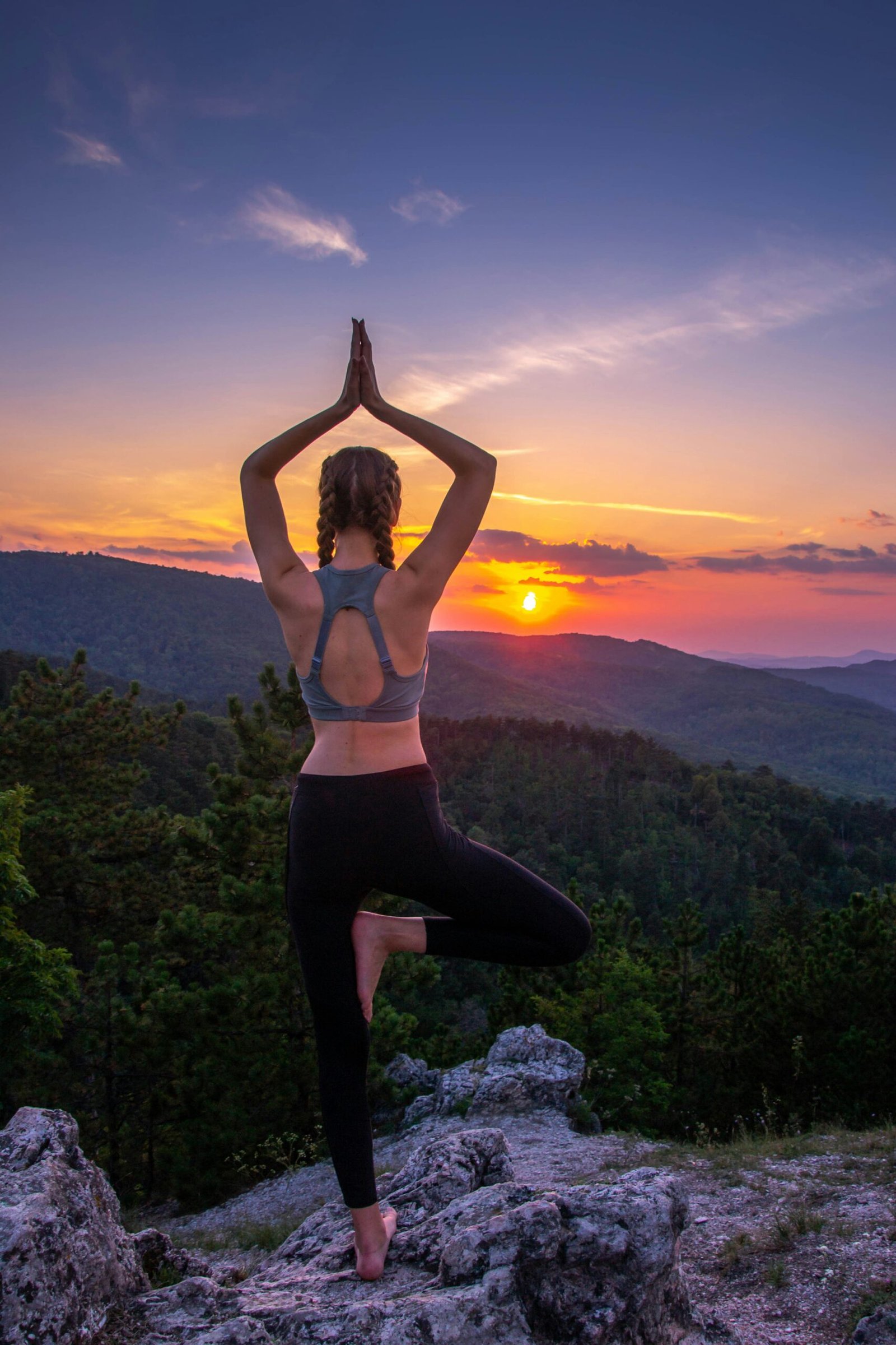 A woman practicing a yoga tree pose on a mountain summit at sunset, symbolizing the balance and renewal found during an immersive yoga retreat.