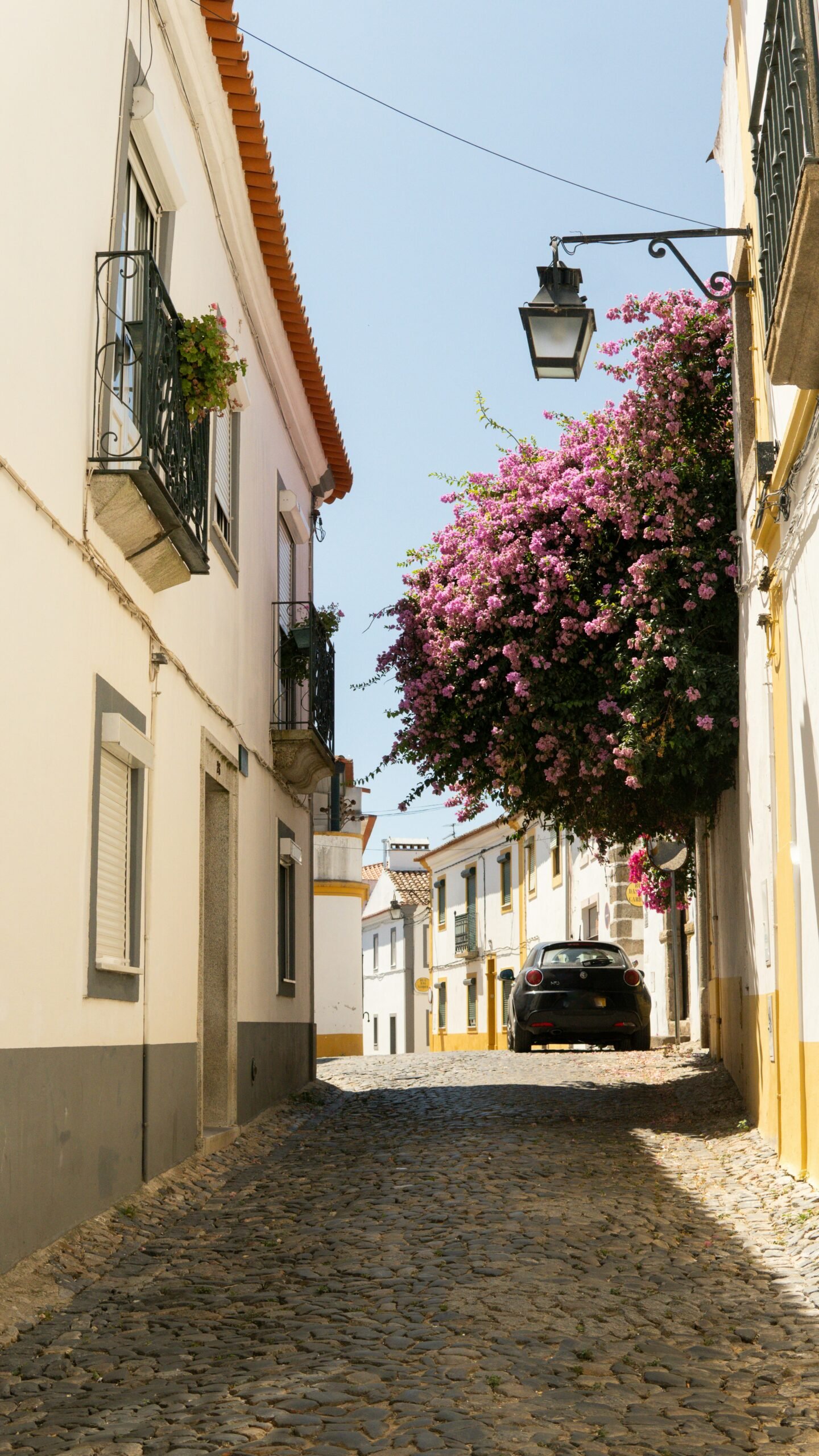 A narrow cobblestone street in a traditional Portuguese village featuring white buildings with yellow trim and vibrant pink bougainvillea, perfect for a peaceful spring yoga retreat.