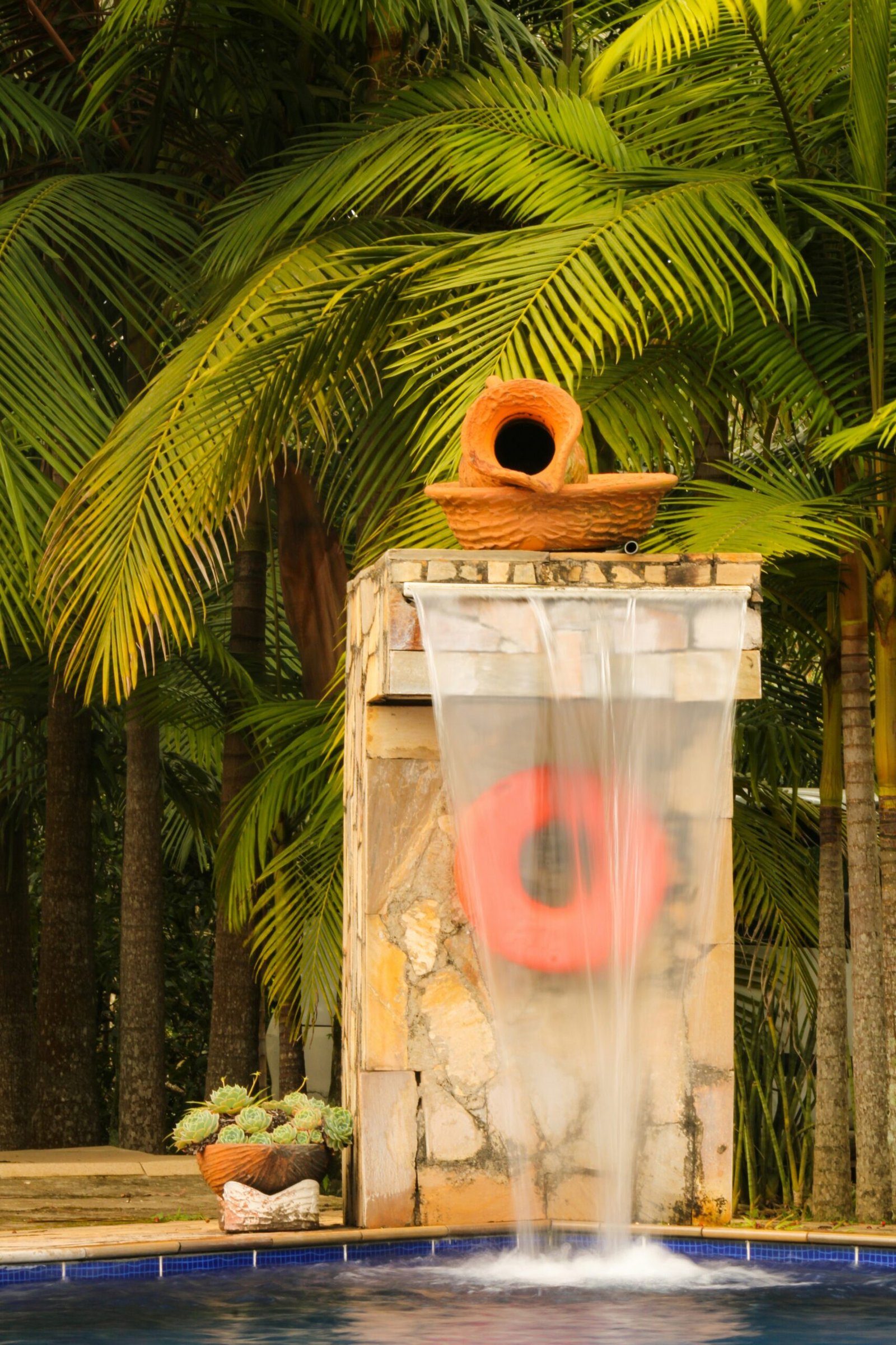 Tropical water feature with a terracotta pot and life preserver in a resort-style pool setting.
