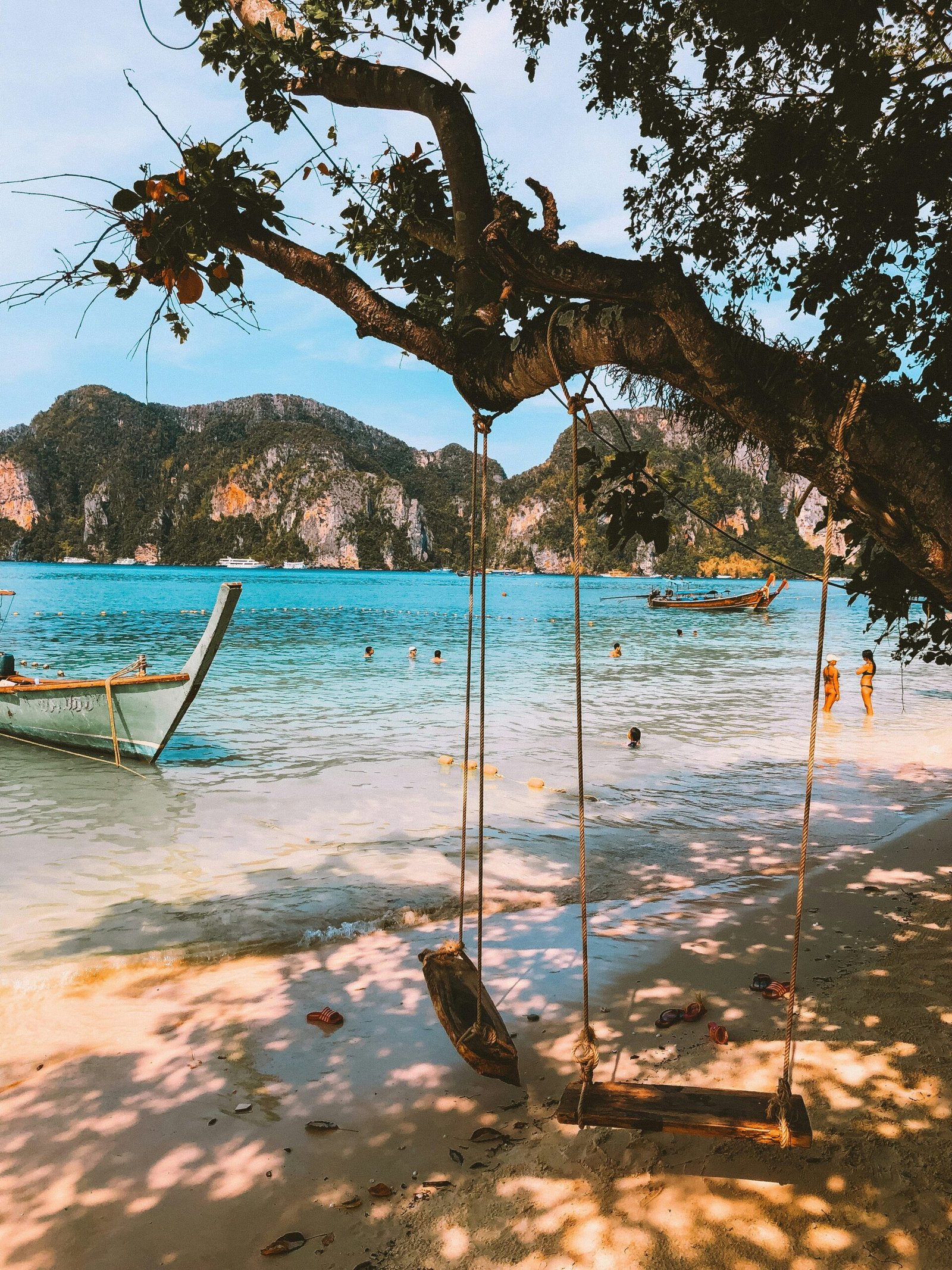 A rustic wooden swing on a white sand beach in the Phi Phi Islands, Thailand, featuring limestone cliffs and a traditional longtail boat for a 2026 yoga retreat guide.