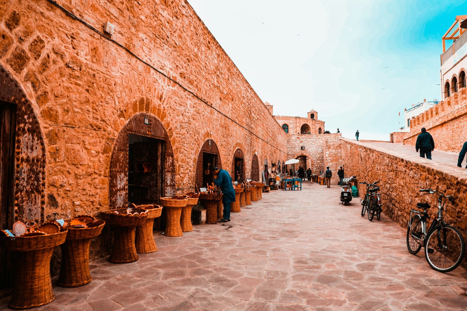 "A stone-walled market alley in the historic coastal city of Essaouira, Morocco, featuring traditional artisanal baskets and local crafts under a bright summer sky.