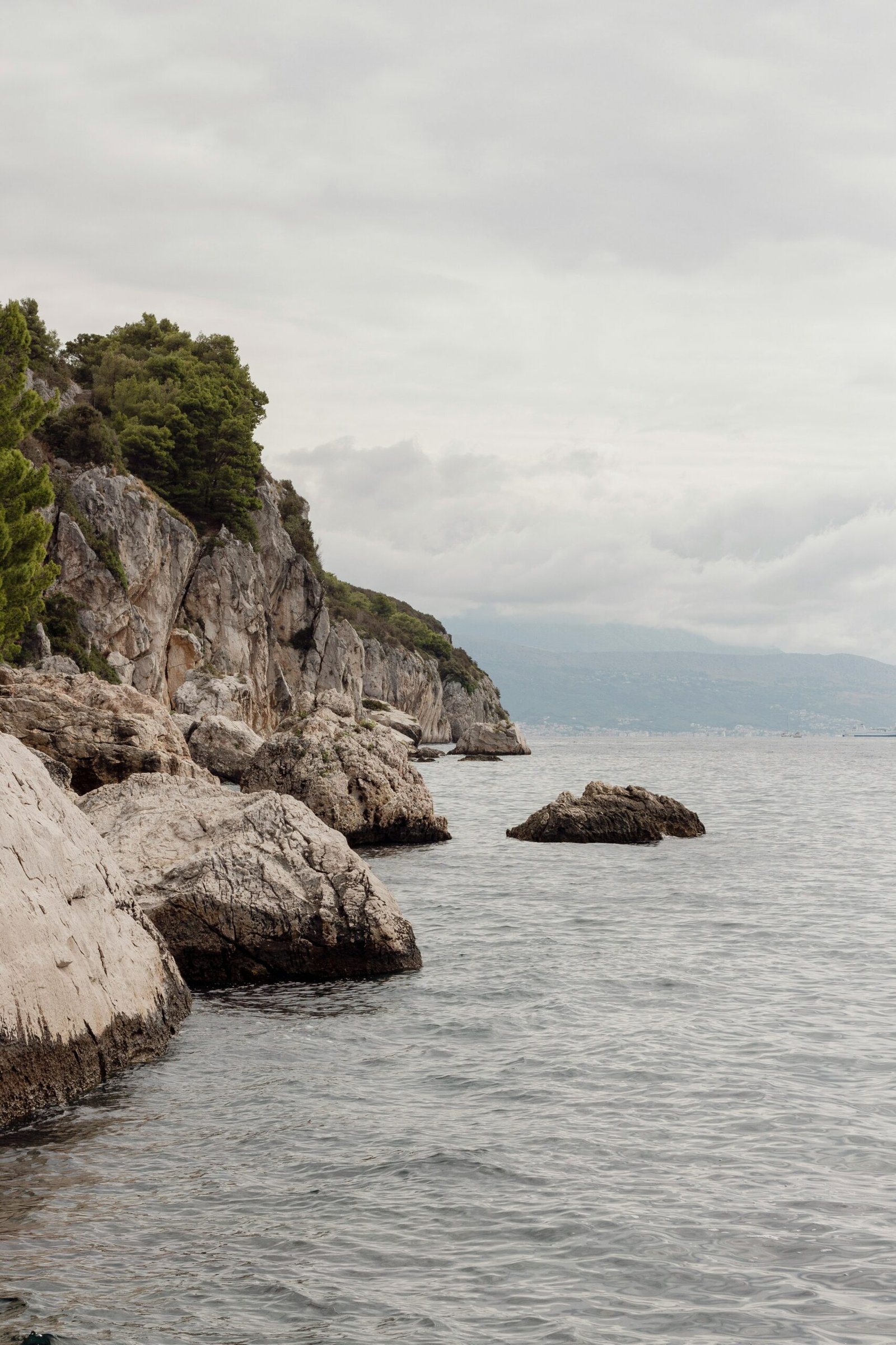 A serene rocky coastline with calm grey waters and lush green cliffs, illustrating the tranquil and reflective environment of a coastal yoga retreat in Spain.