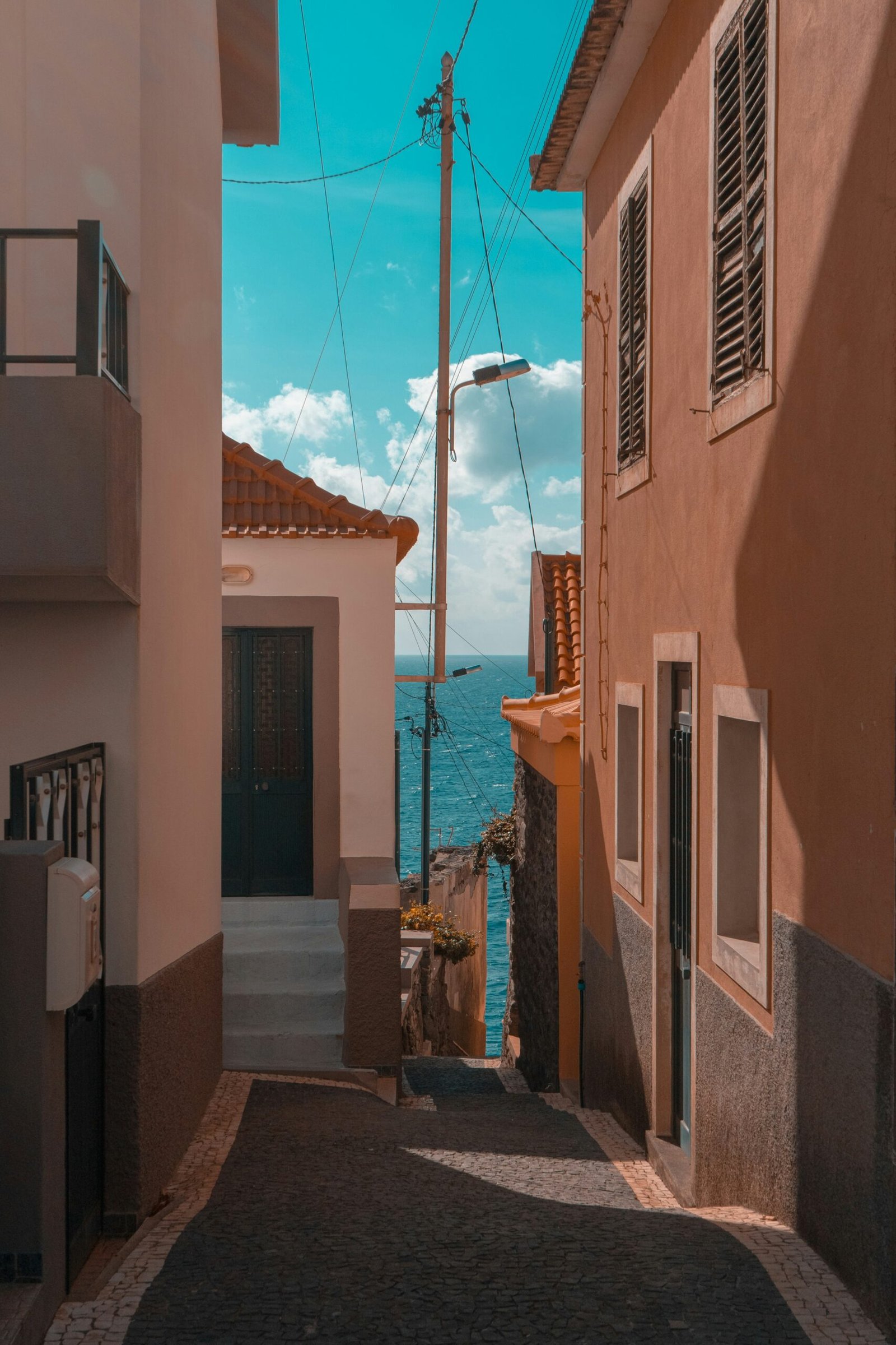 A narrow, sunlit street in Madeira, Portugal, with traditional orange-toned houses leading toward a deep blue ocean view, representing a serene location for a winter yoga retreat.