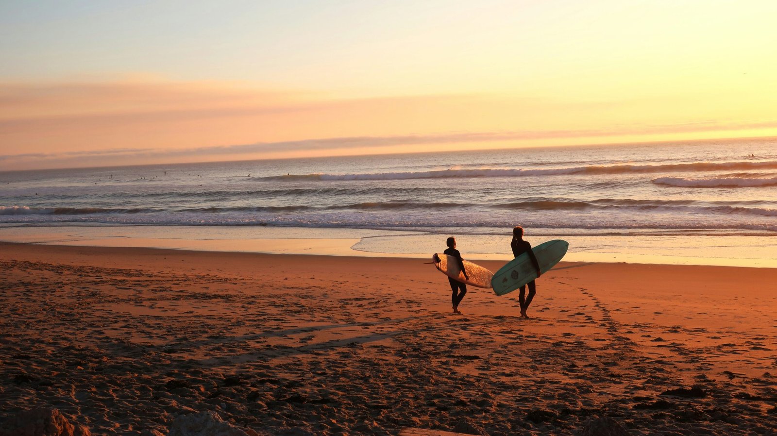Two surfers carrying boards along a golden beach at sunset in Portugal, highlighting the popular surf and yoga retreat season during the warm autumn months.