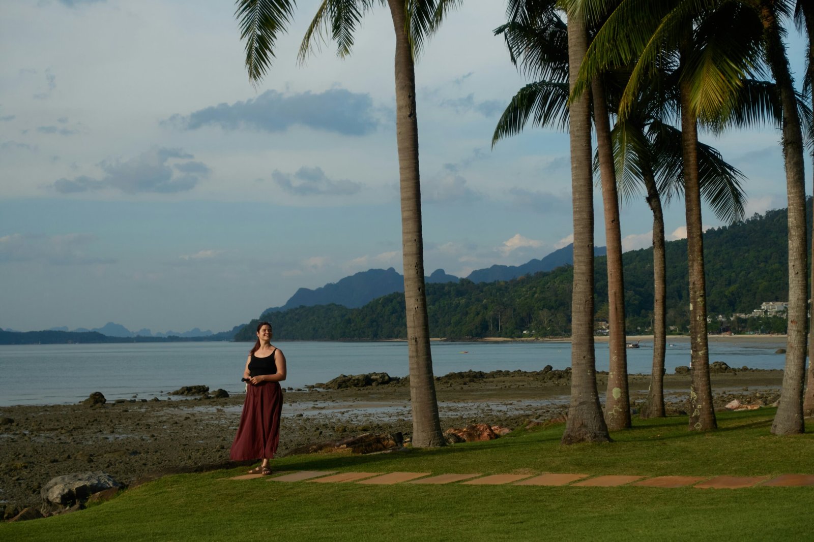 A woman smiling on a manicured lawn lined with palm trees overlooking a tropical bay, representing the premium value of high-end resort grounds at an all-inclusive yoga retreat.