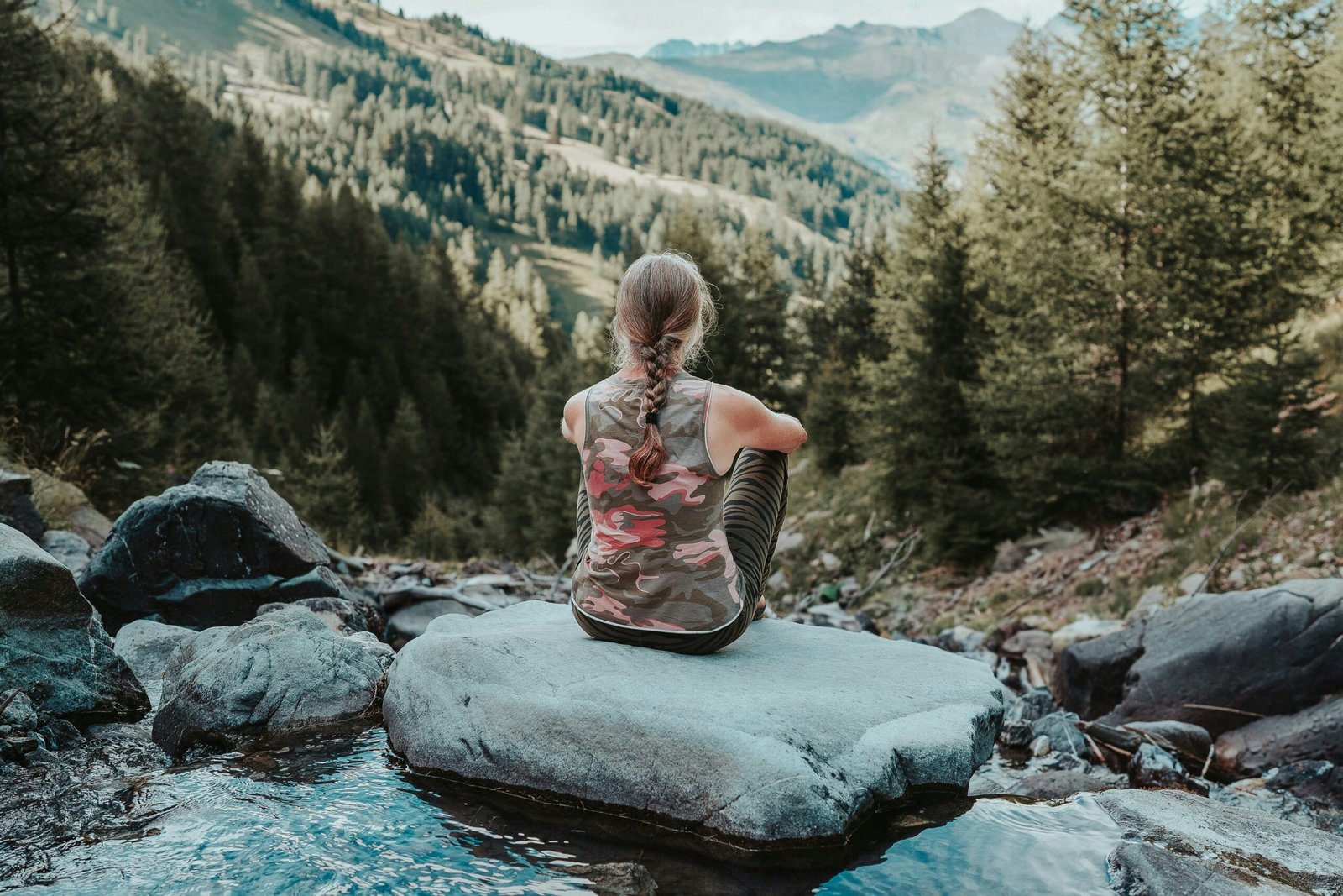 A woman sitting in a meditative pose on a large rock by a mountain stream, representing the mental clarity and inner peace achieved through a nature-based yoga retreat.
