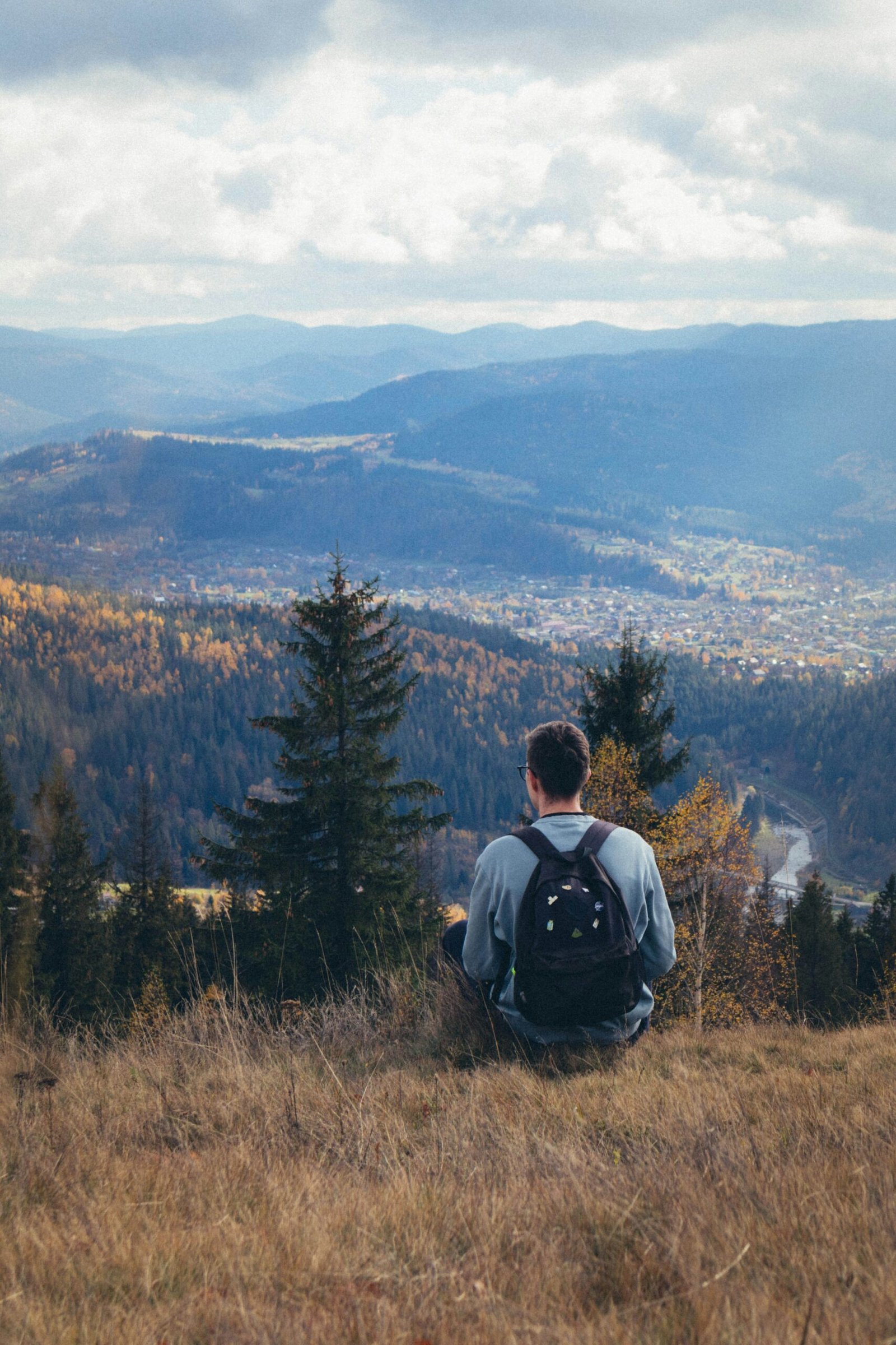 A lone hiker with a backpack sits on a grassy hill, overlooking a vast mountain valley and village, demonstrating the peaceful feeling of recentering in nature through daily breathwork.