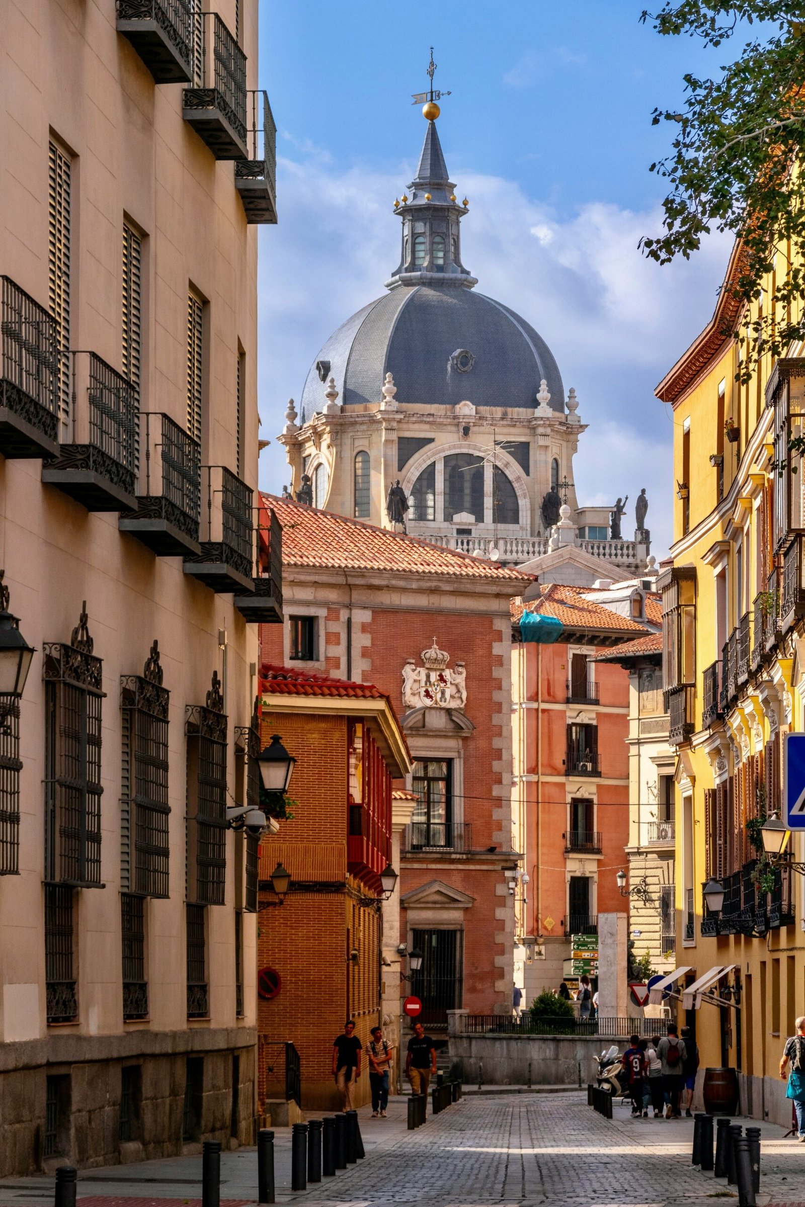 A narrow historic street in Madrid leading toward the grand dome of the Almudena Cathedral under a blue sky, representing the architectural beauty of the city before traveling to a quiet yoga retreat.