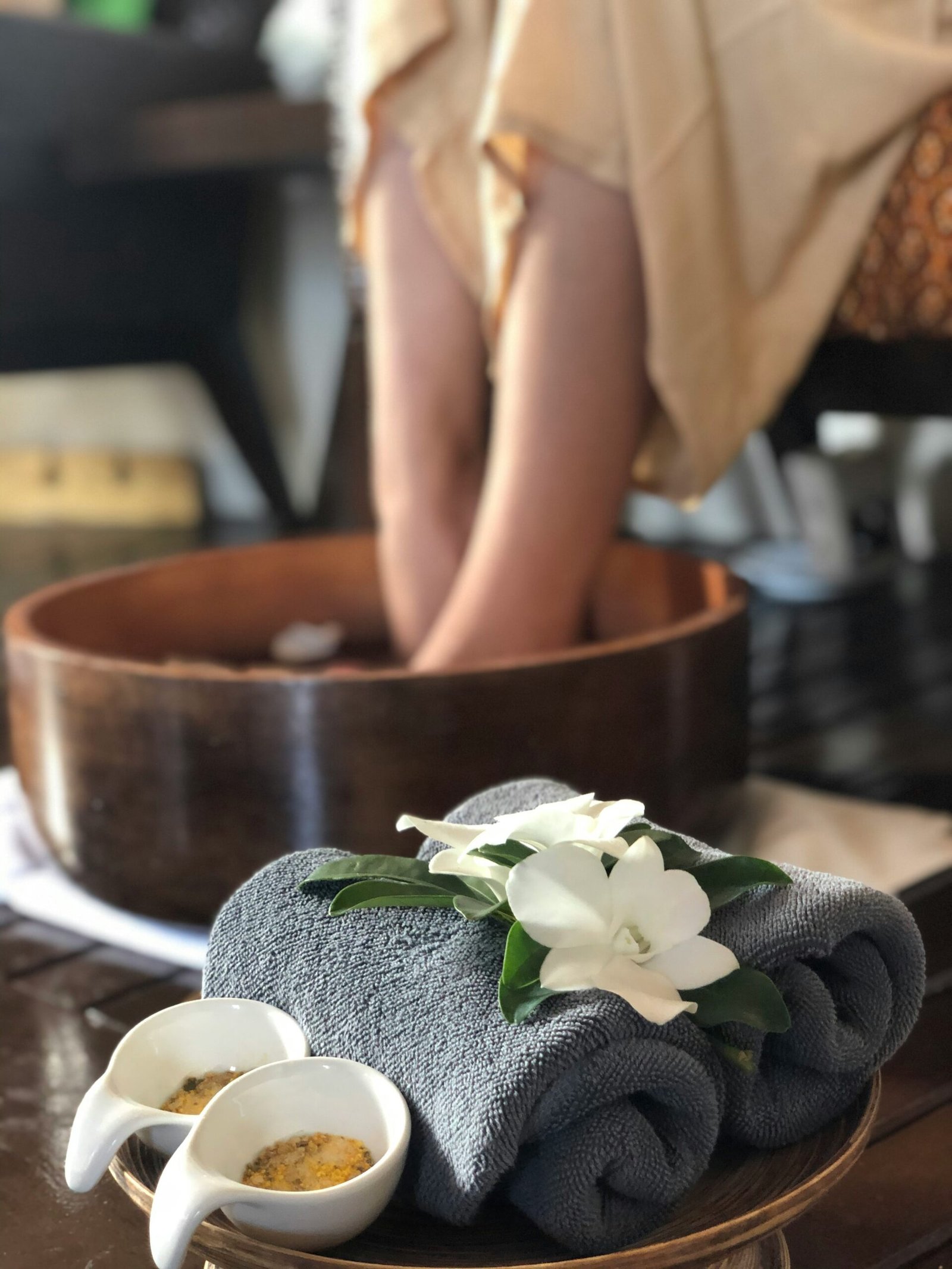 erson soaking both feet in a wooden bowl during a calming spa-style foot ritual with flowers nearby.