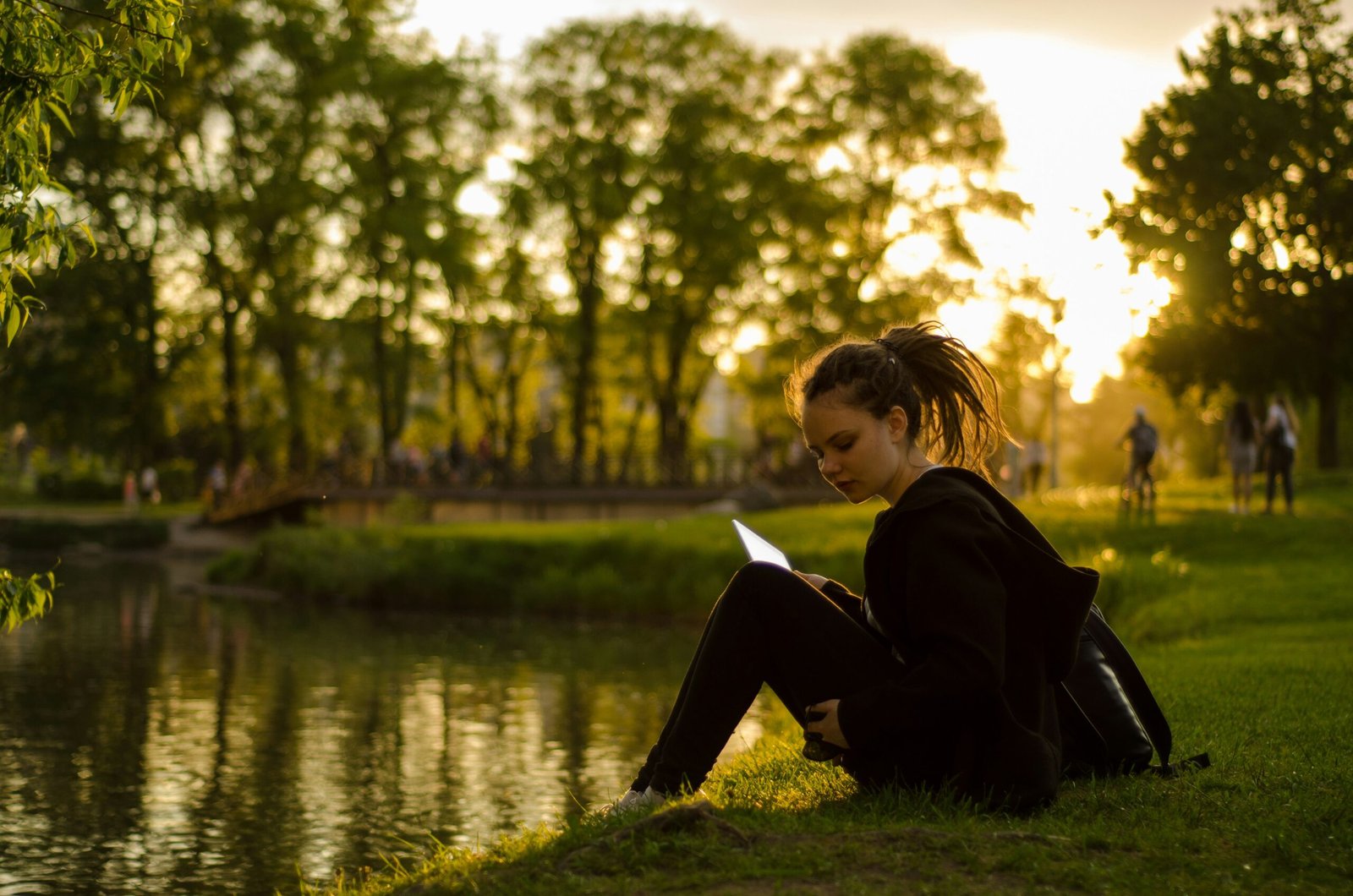 A woman looking at a screen by a lake at sunset, representing the digital fatigue that prompts travelers to seek a restorative digital detox retreat.