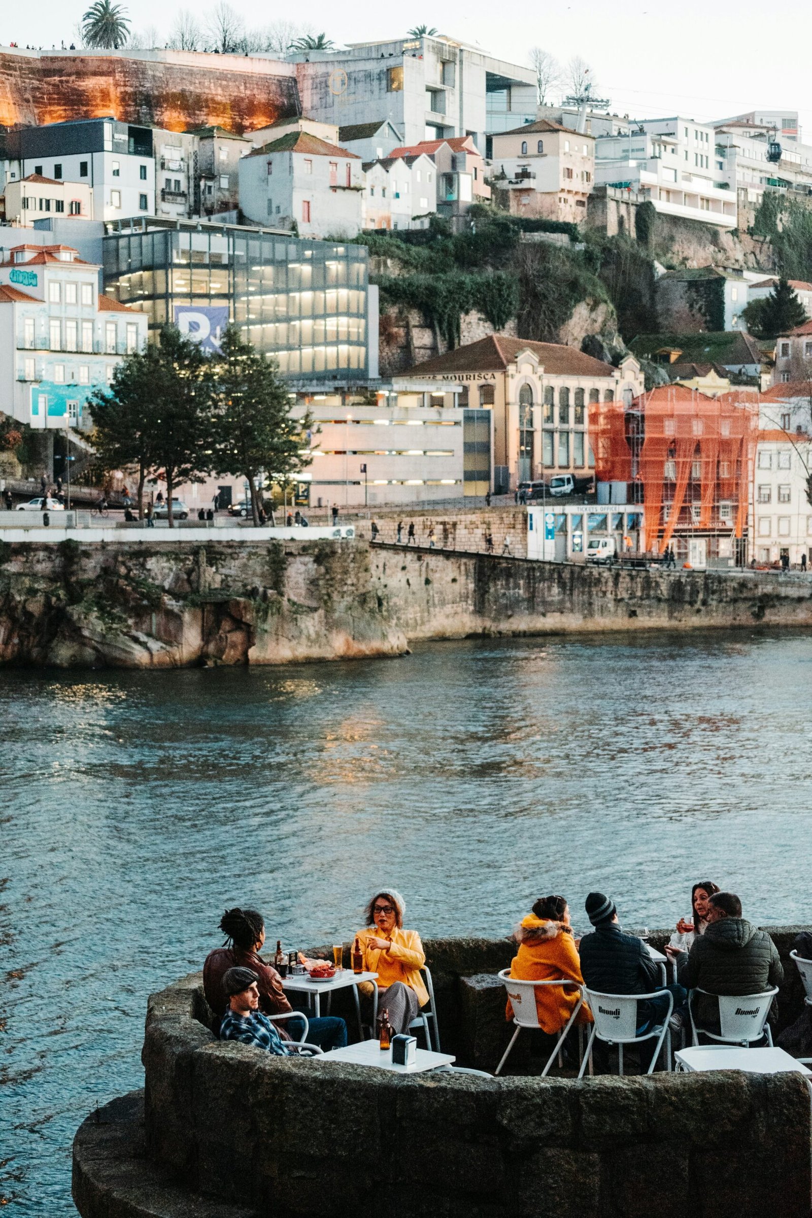 A group of people enjoying drinks at an outdoor riverside café in Porto, Portugal, during winter, showcasing the social and cozy 'slow travel' lifestyle of a wellness retreat.