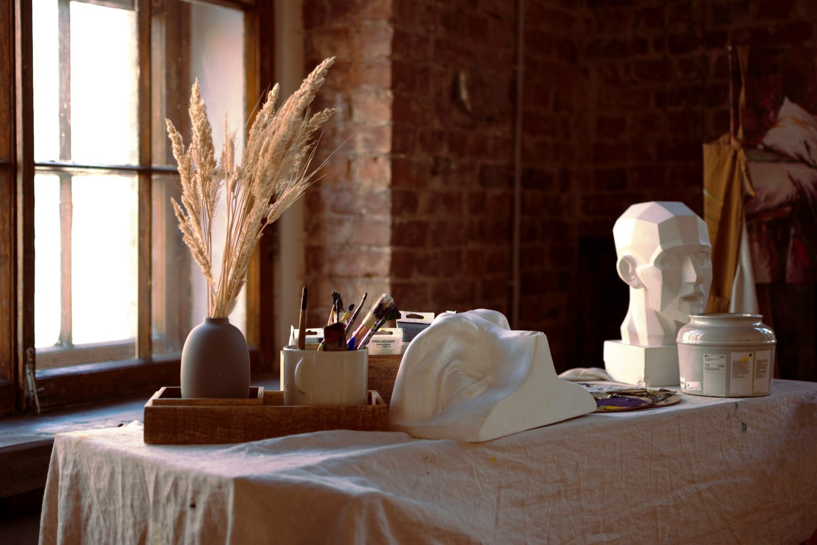 An artist’s studio table with sculptures, brushes, and dried wheat by a sunlit window, representing the creative expression and art therapy workshops common in holistic wellness retreats.