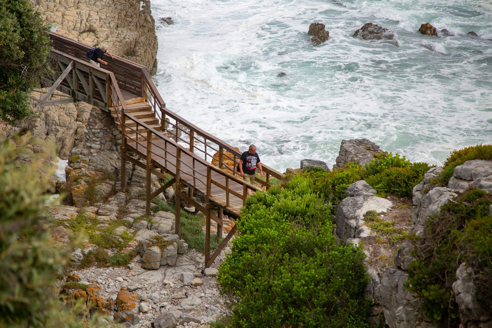 A person walking down a wooden boardwalk path built into the rugged limestone cliffs of the Algarve coastline, illustrating slow travel and mindful hiking near Lagos, Portugal.