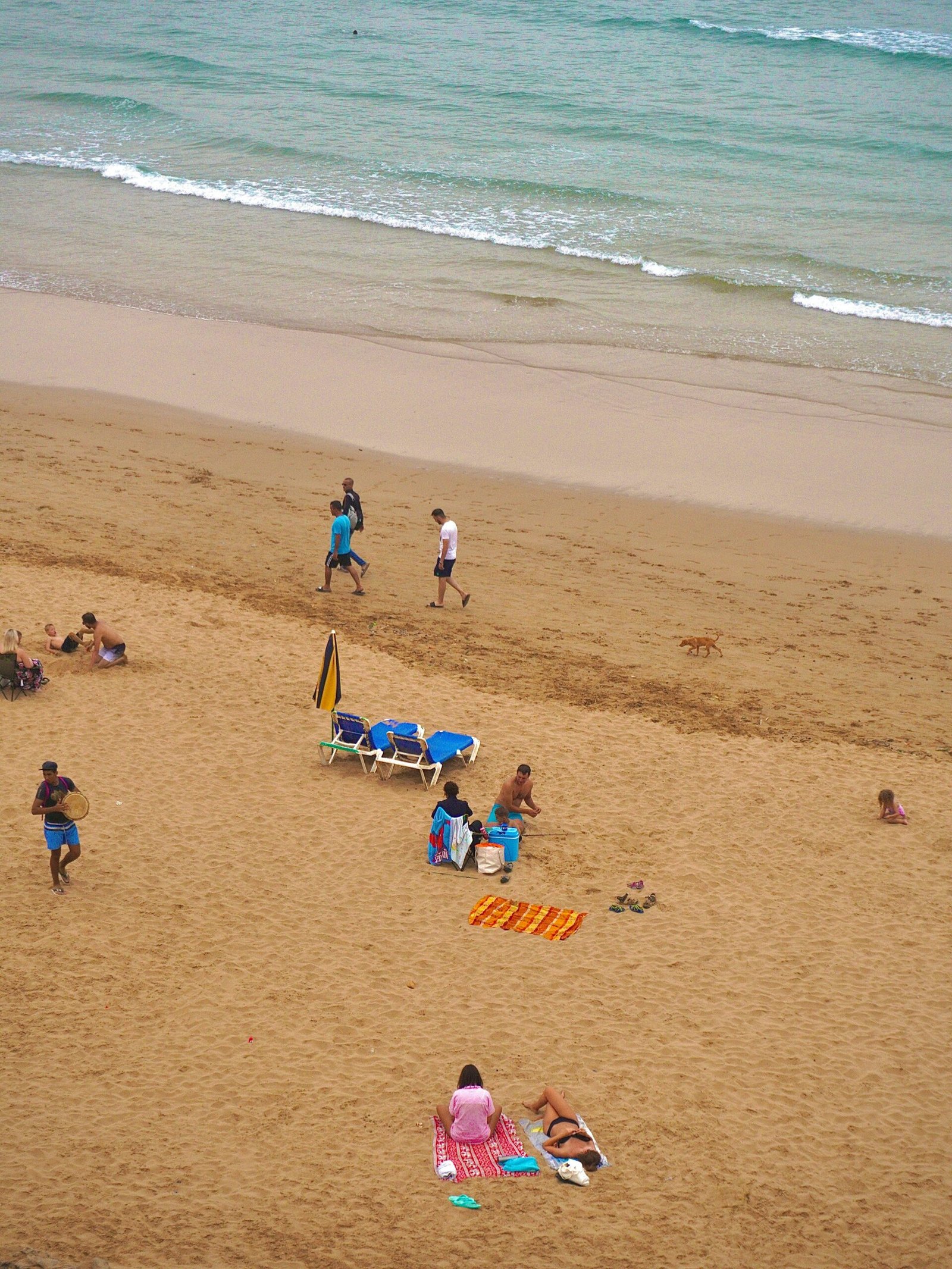 An overhead view of people relaxing and walking on a wide sandy beach in Morocco with gentle Atlantic waves, perfect for a summer yoga and social wellness retreat.